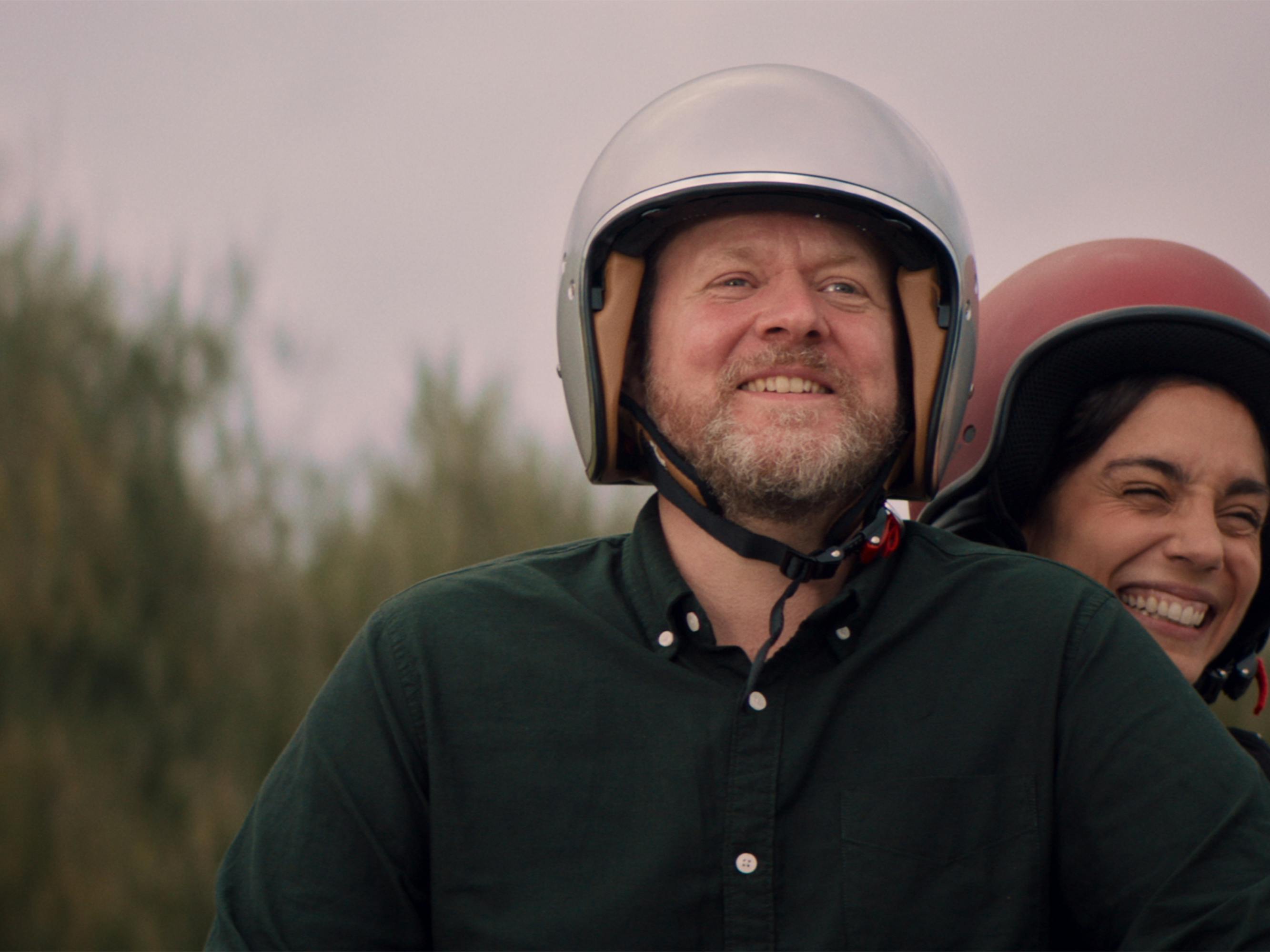 Theo (Anders Matthesen) and Sophia (Cristiana Dell’Anna) ride on a motorcycle wearing white and red helmets respectively, looking elated. 