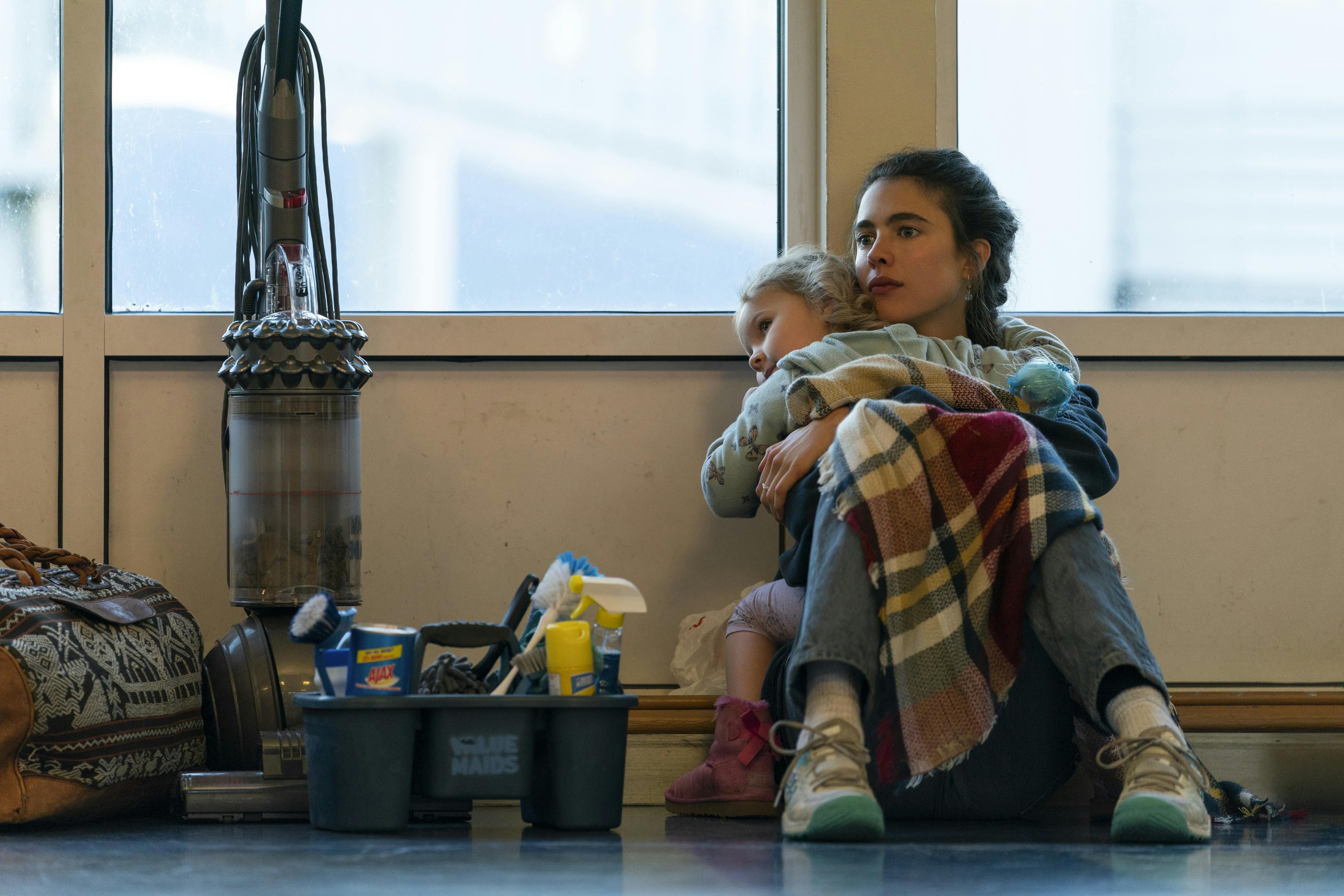 Alex Russell (Margaret Qualley) and Maddy (Rylea Nevaeh Whittet) sit amongst cleaning supplies on the floor of a starkly-lit room.