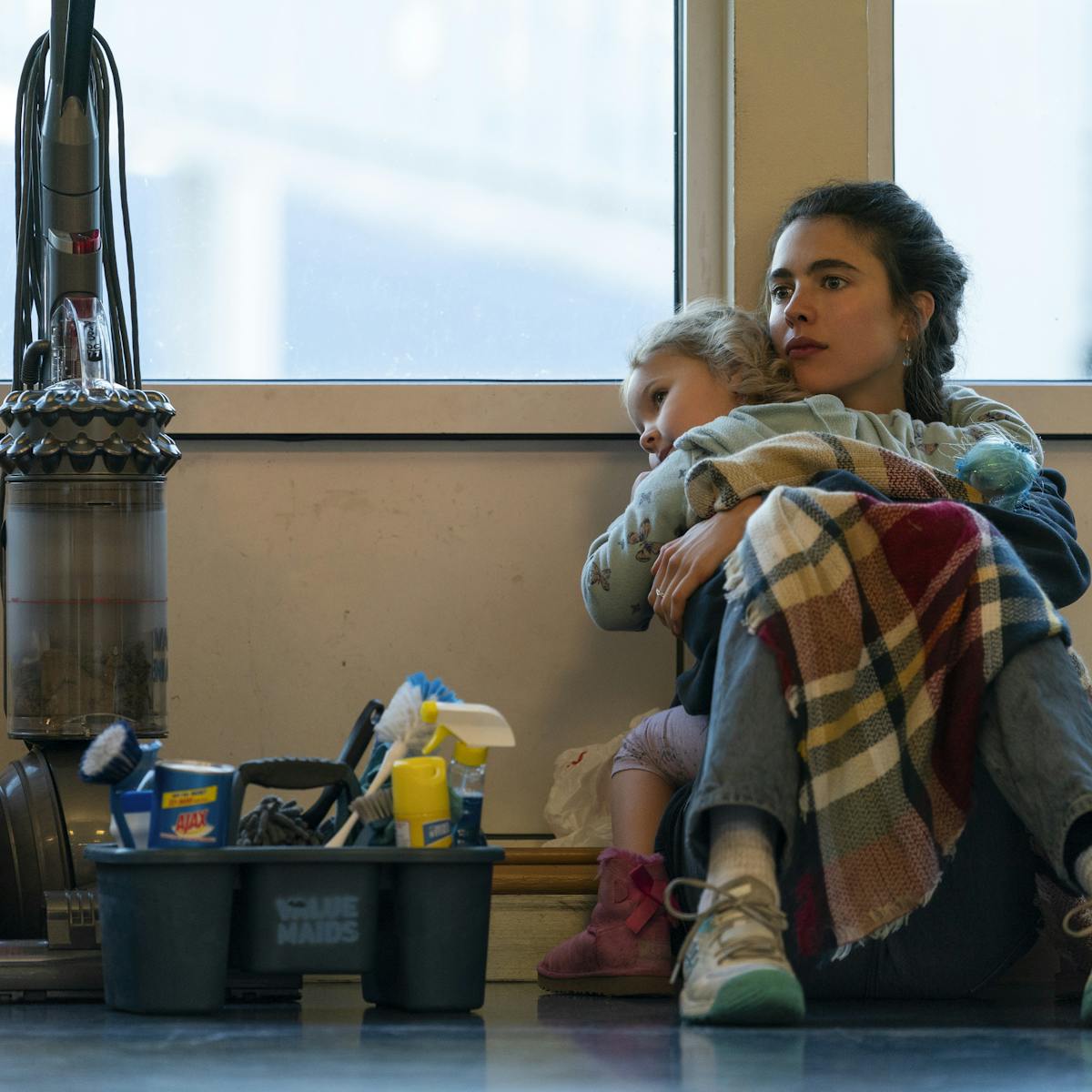 Alex Russell (Margaret Qualley) and Maddy (Rylea Nevaeh Whittet) sit amongst cleaning supplies on the floor of a starkly-lit room.