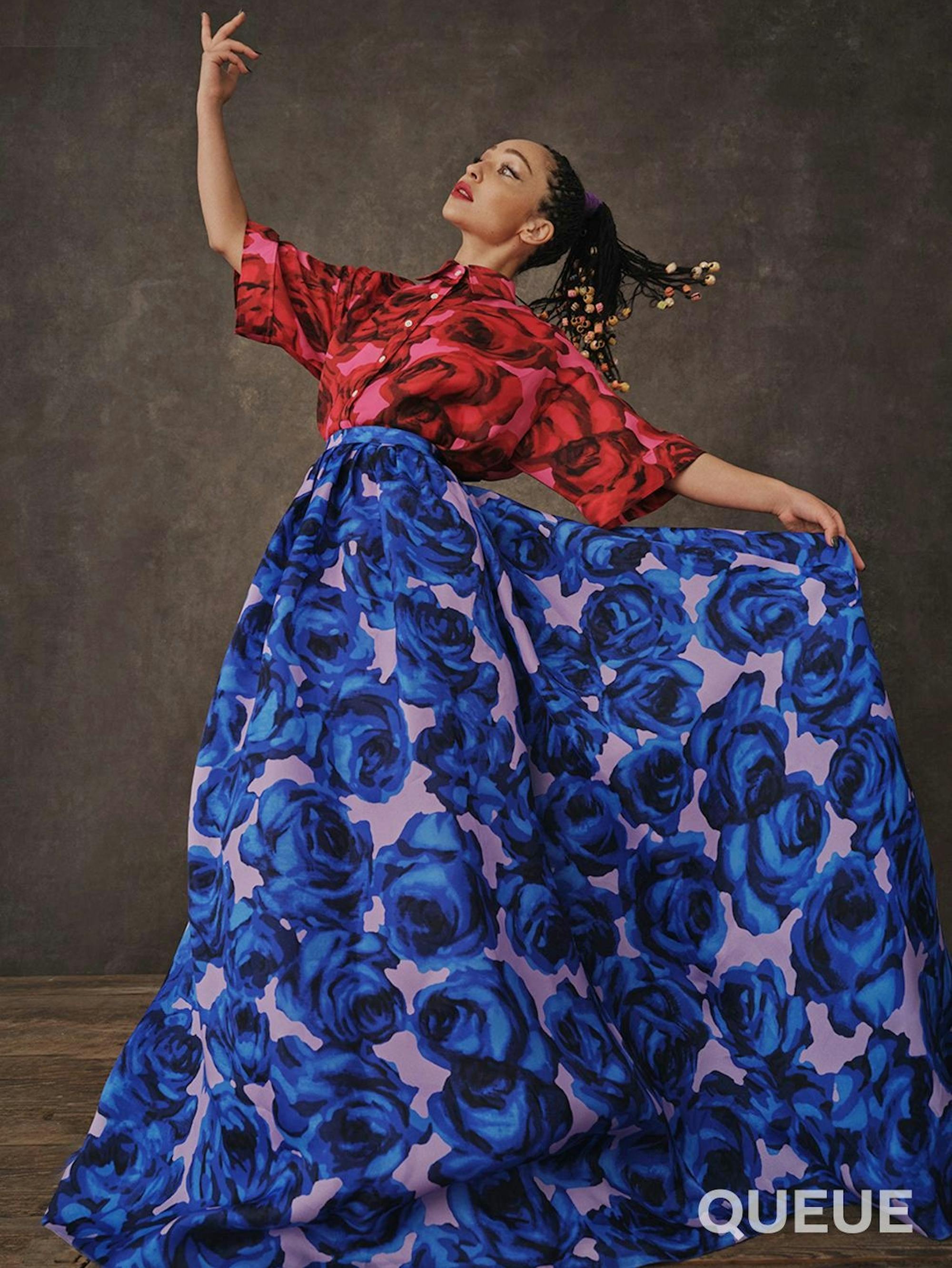 Ruth Negga wears a purple skirt with blue and black flowers, and a pink shirt with red and black flowers. They’re both shot mid dance move, giving these images movement and energy!