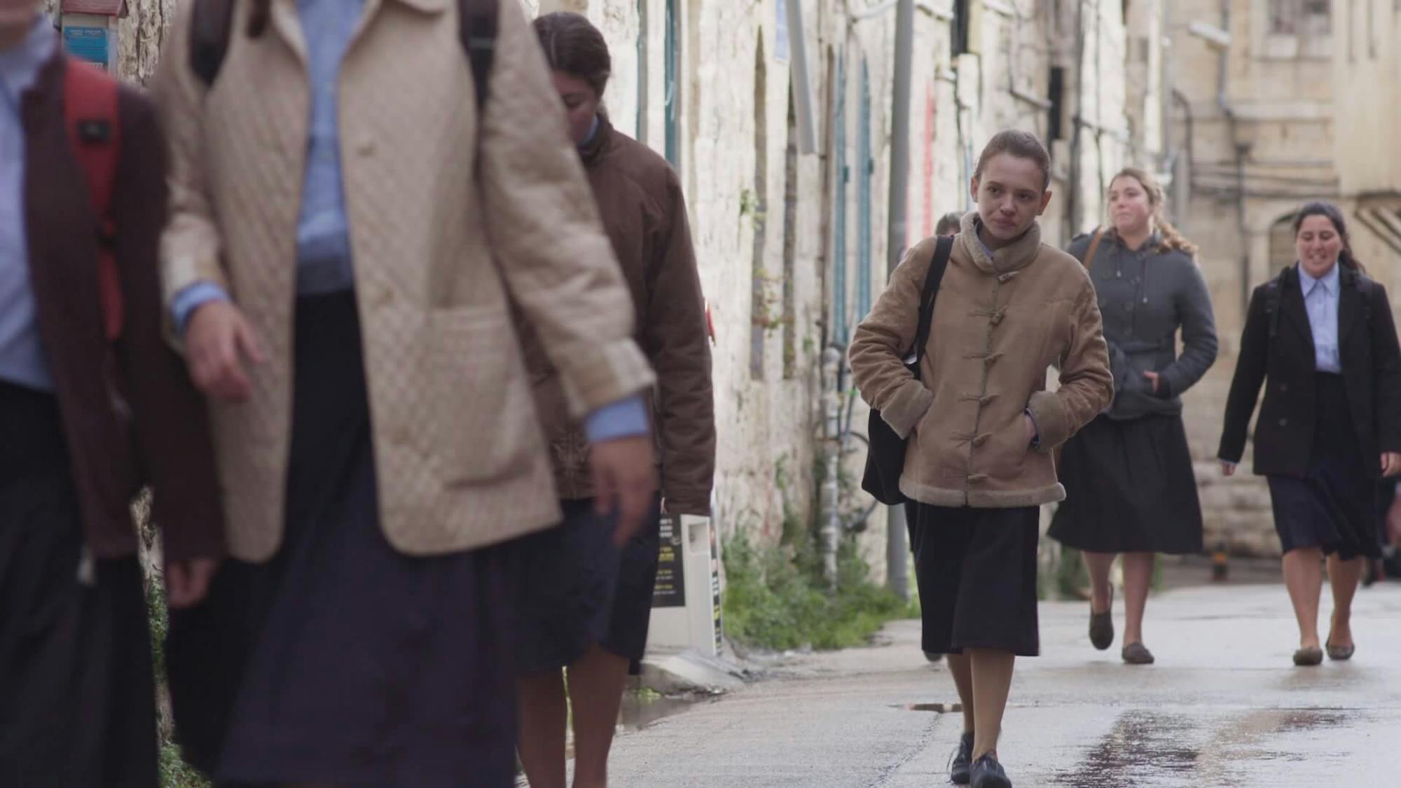 Ruchama Weiss (Shira Haas) walks down the street with others dressed in similar colors and outfits: black, navy skirts, light blue buttoned-down shirts, and tan/brown jackets.