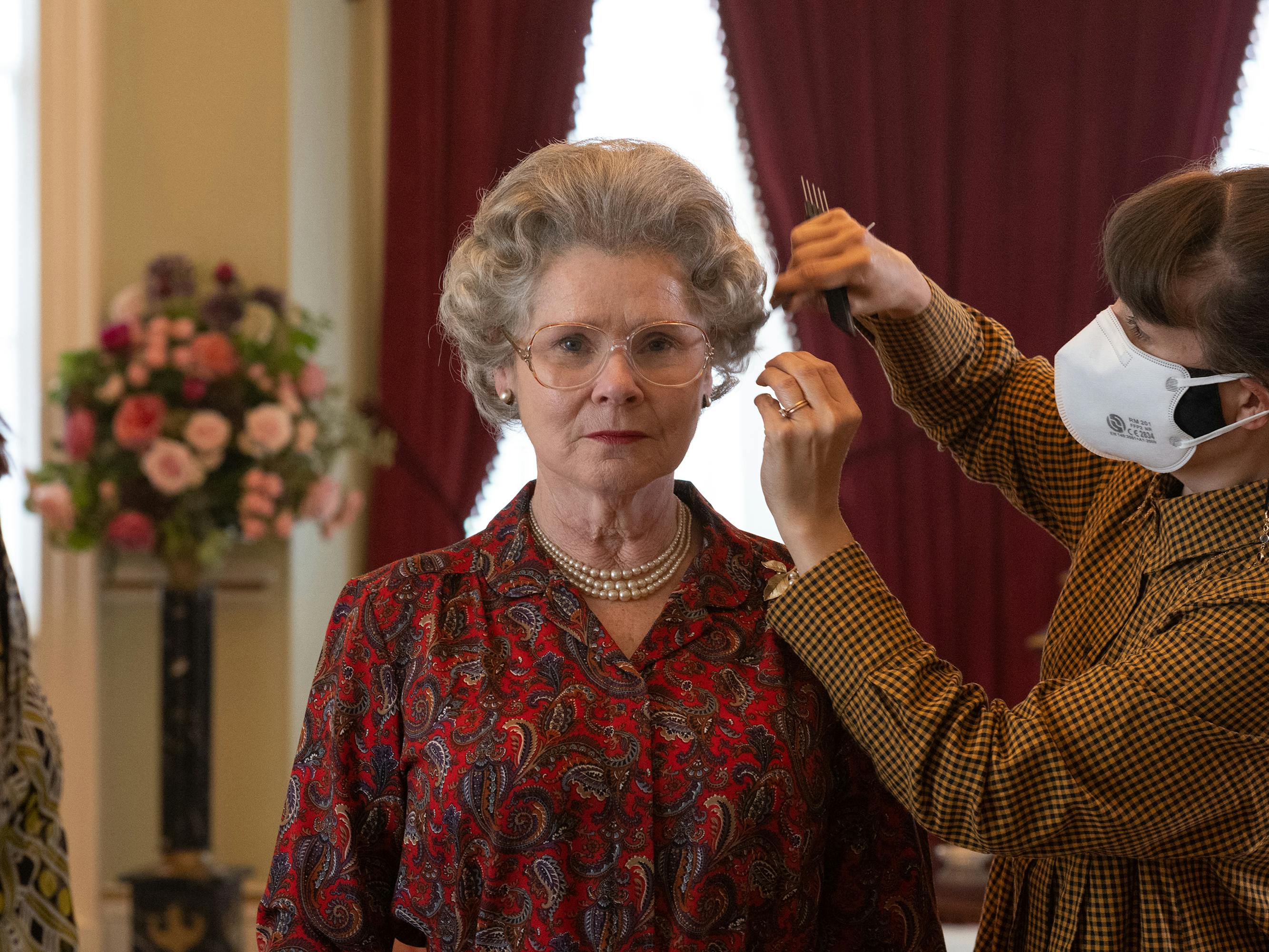 Imelda Staunton gets her hair touched up by someone in a mask. Staunton wears a red patterned shirt and looks straight ahead.