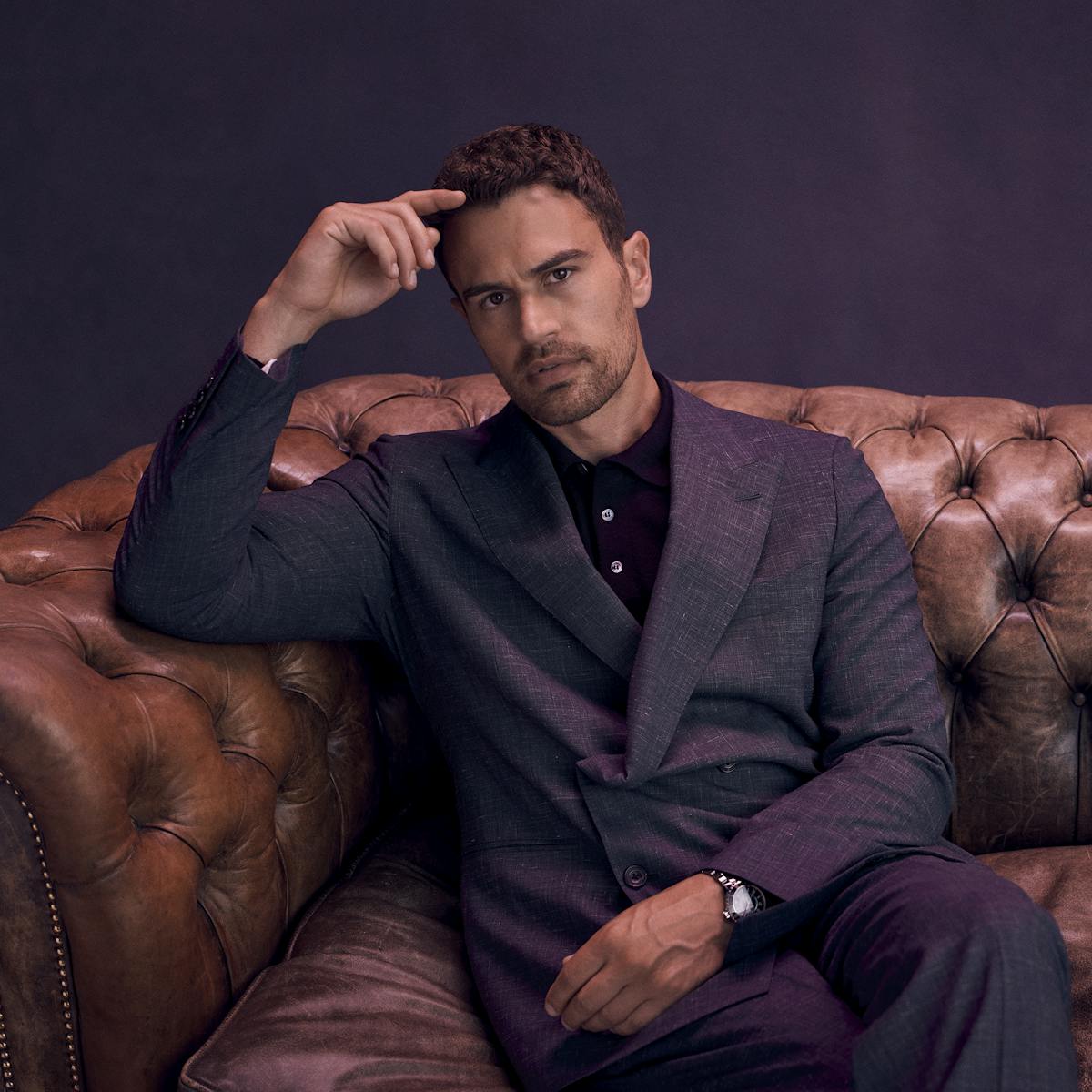 Theo James wears a purple suit and sits on a brown leather couch.