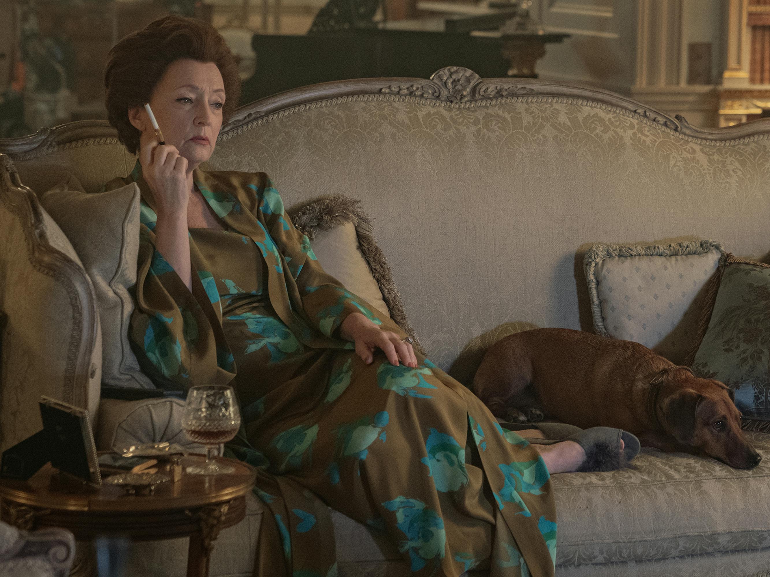 Princess Margaret (Lesley Manville) wears a patterned dress and sits on a couch with a brown dog.