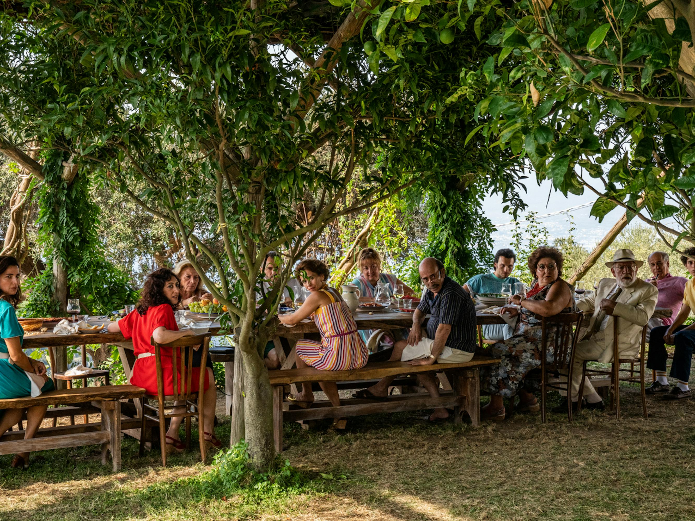 The cast of The Hand of God sits outside under very green trees at a picnic table.