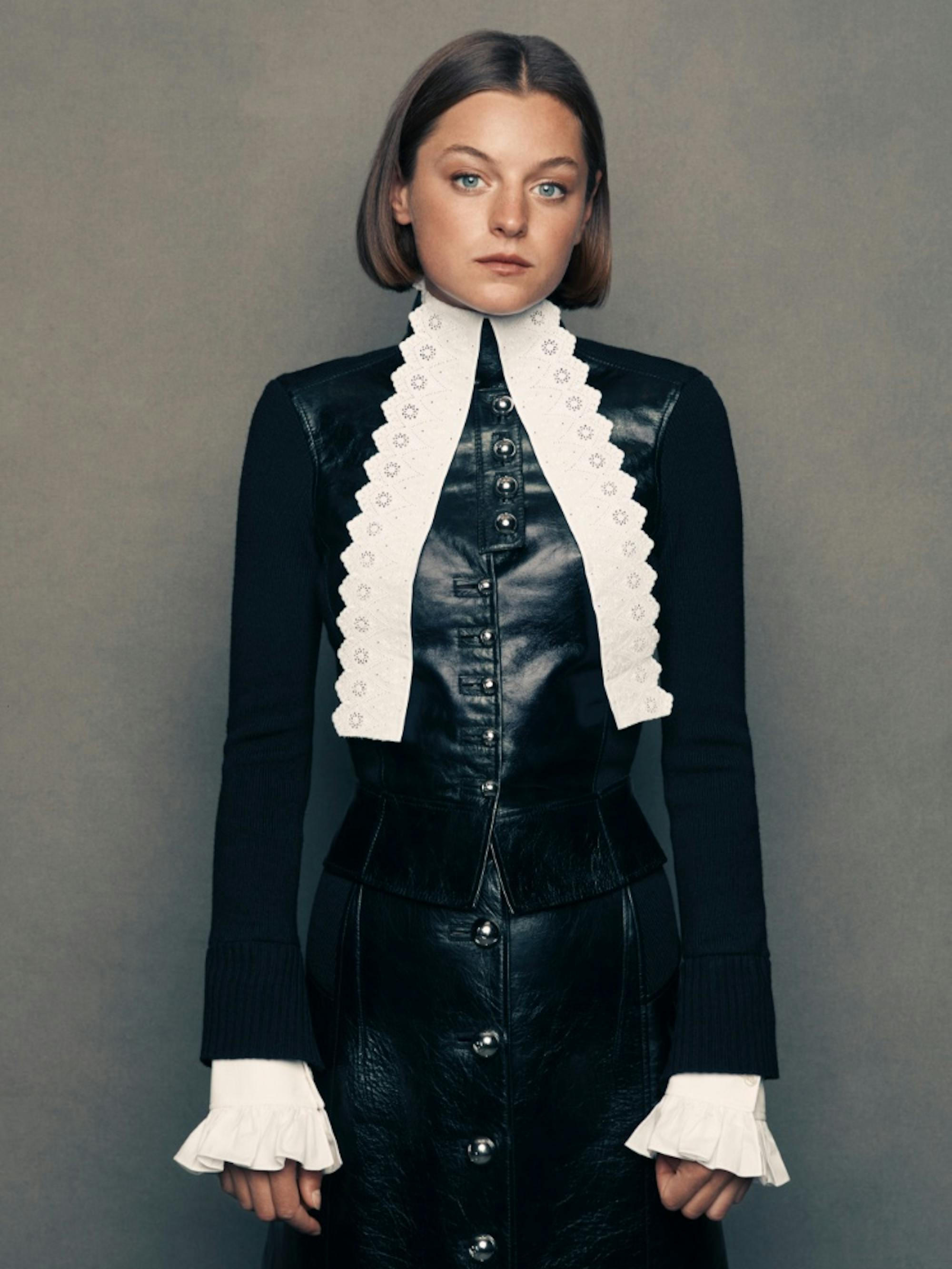 Emma Corrin stands with her hands at her sides, head tilted slightly, in this study of grays, blacks, whites, and browns.