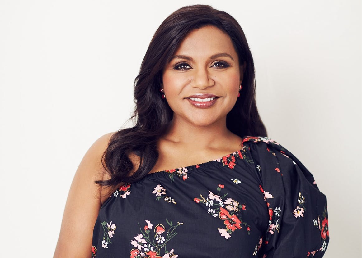 Mindy Kaling wears a black floral, off one shoulder dress. Her hands are crossed at her front, and she smiles.