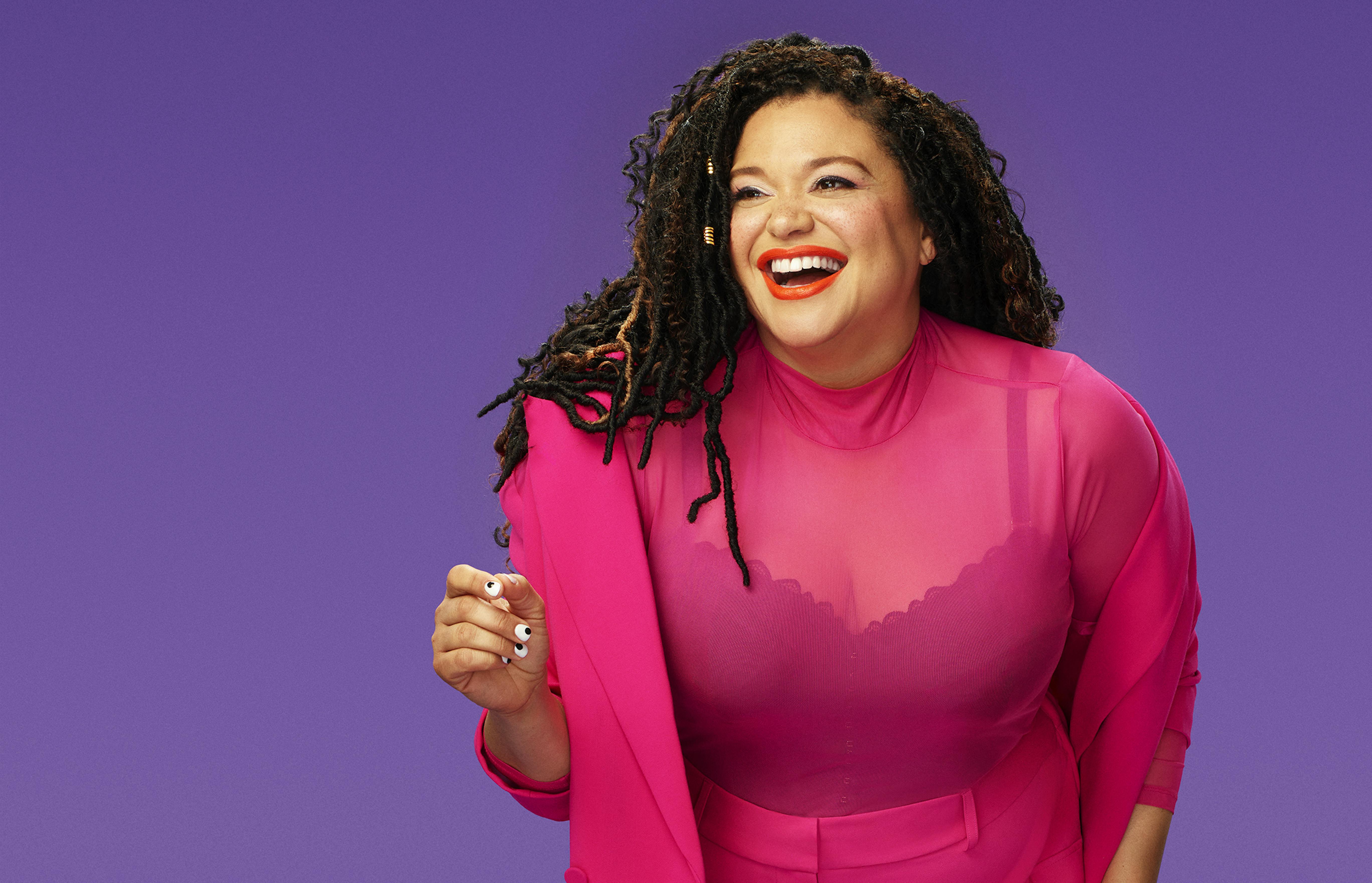 Michelle Buteau wears a pink top over a dark tank, against a purple background.
