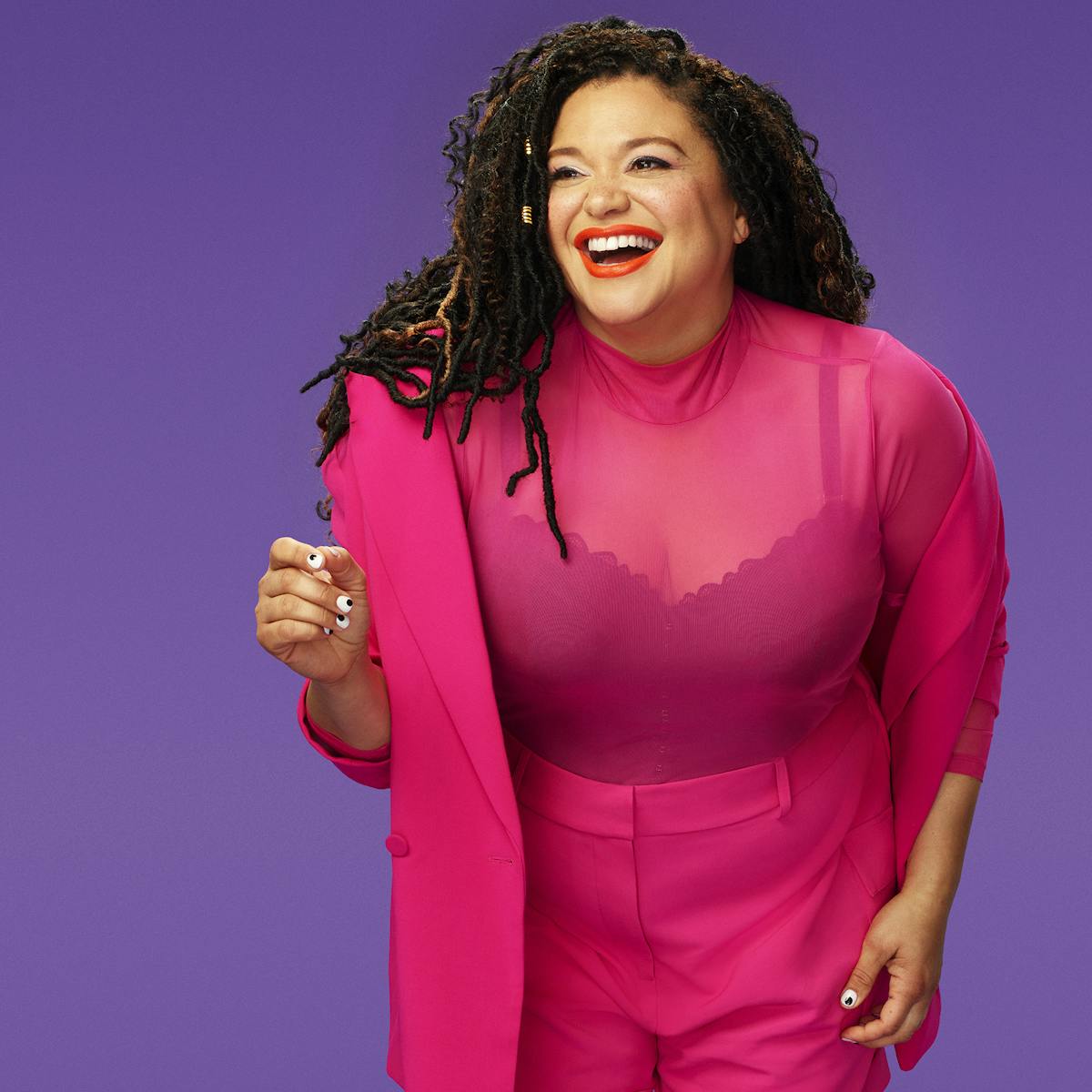 Michelle Buteau wears a pink top over a dark tank, pink shorts, and black heels, against a purple background.