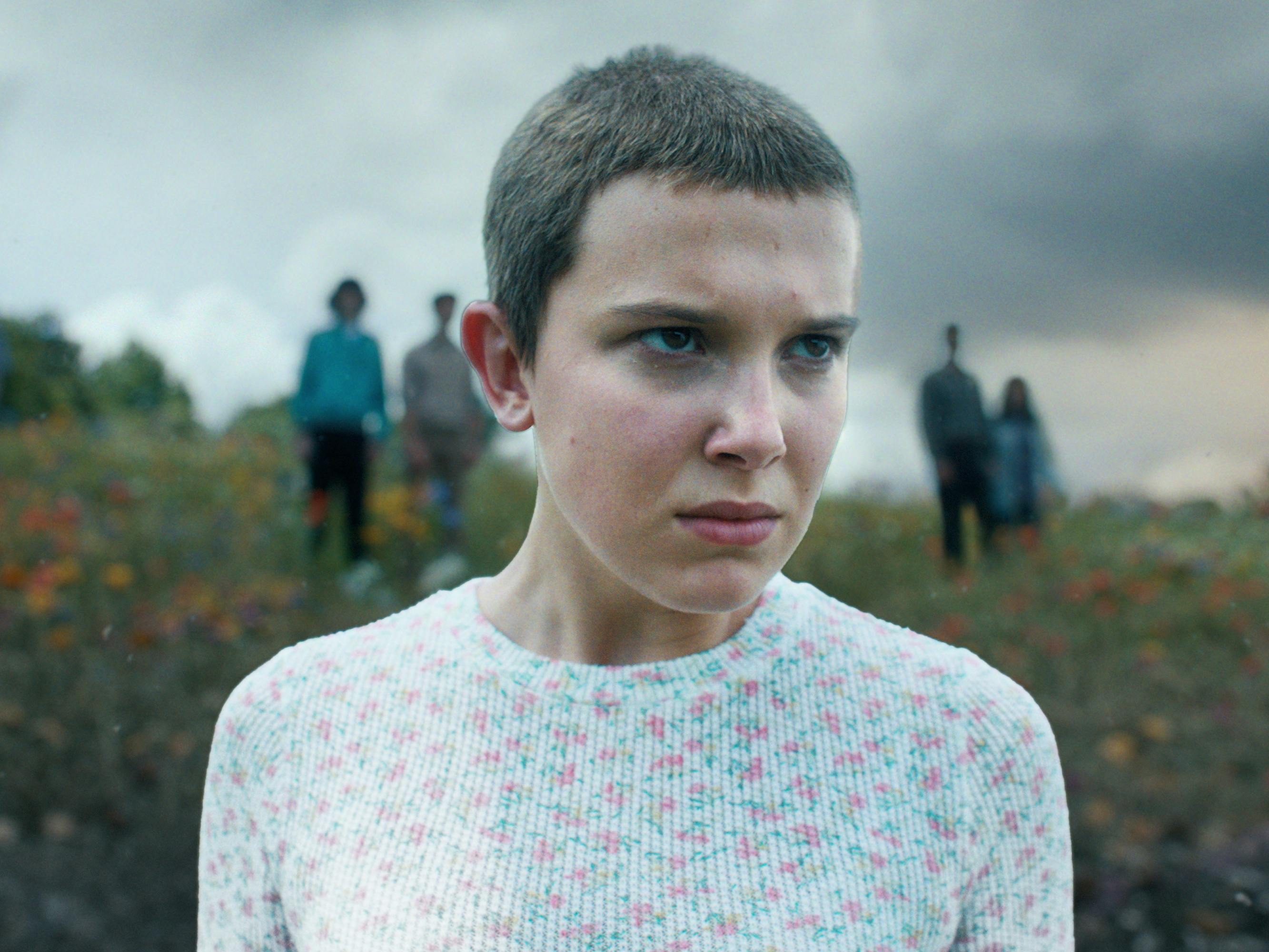 Eleven (Millie Bobby Brown) wears a patterned shirt and stands against a gray sky.