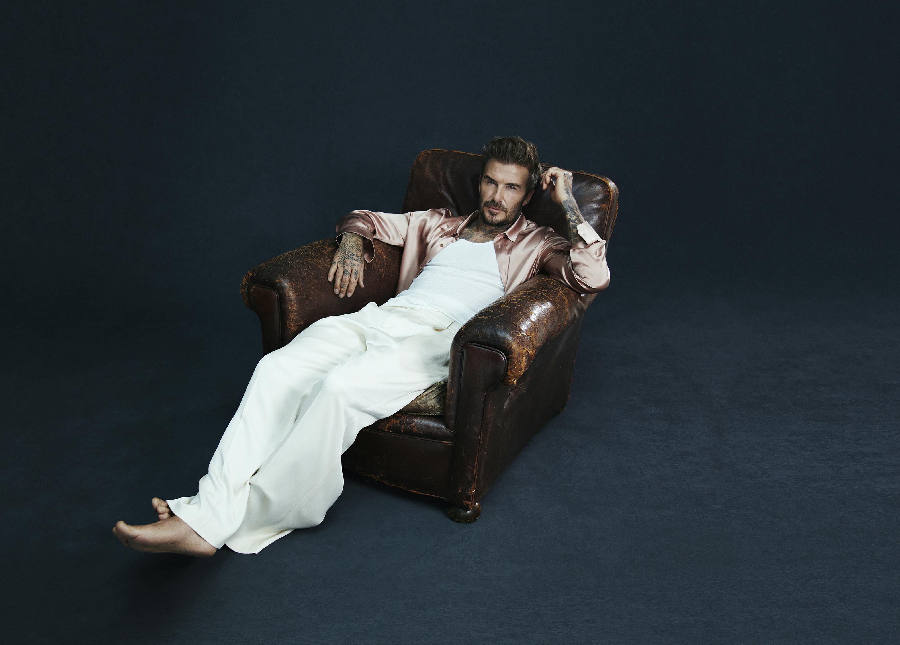 David Beckham wears all white with an open pink silk shirt as he lounges in a brown leather armchair.