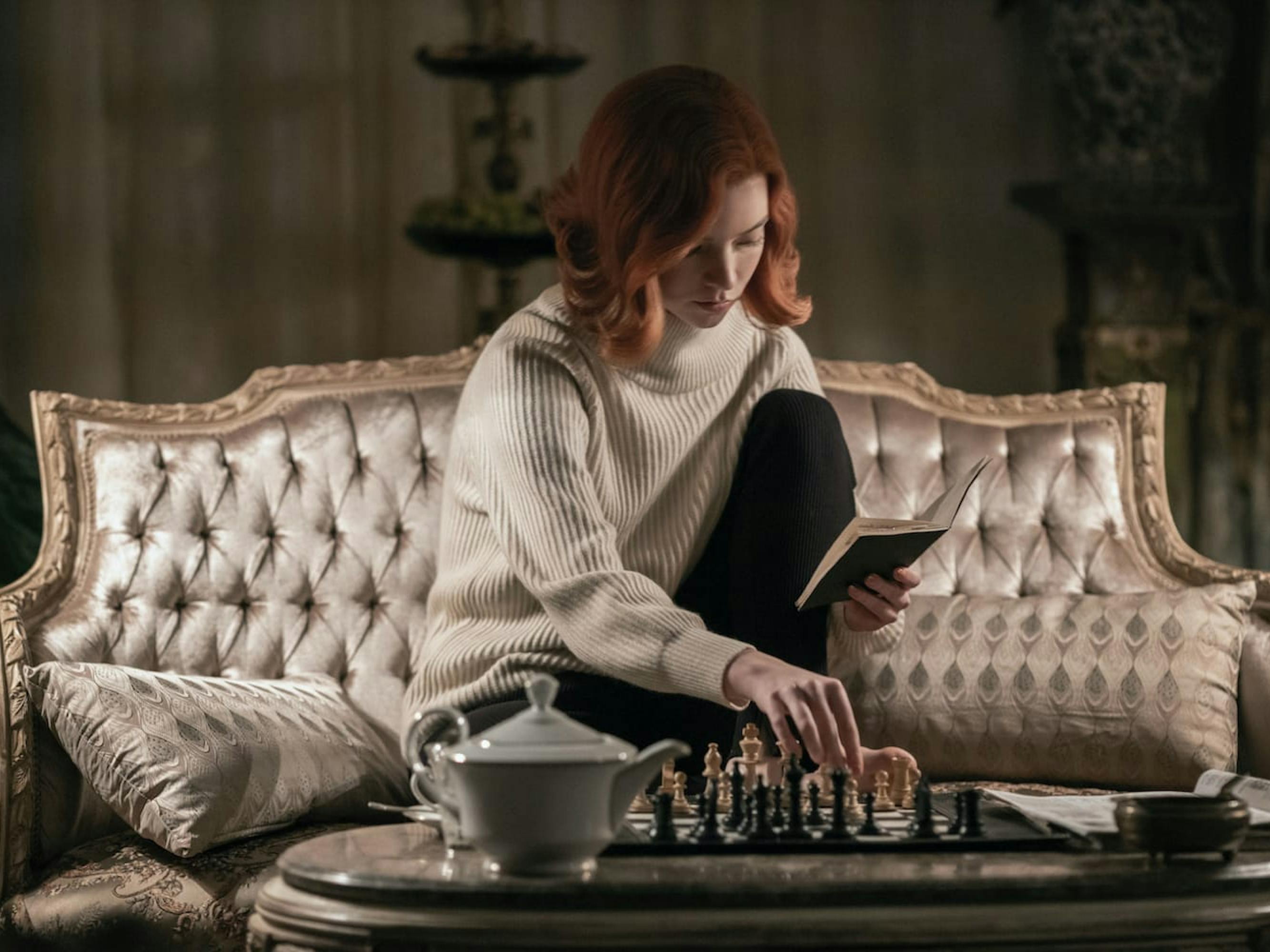 Beth Harmon (Anya Taylor-Joy) plays chess by herself, while reading a small book. She sits on a fabulous, silk, taupe sofa, and enjoys a kettle of tea.