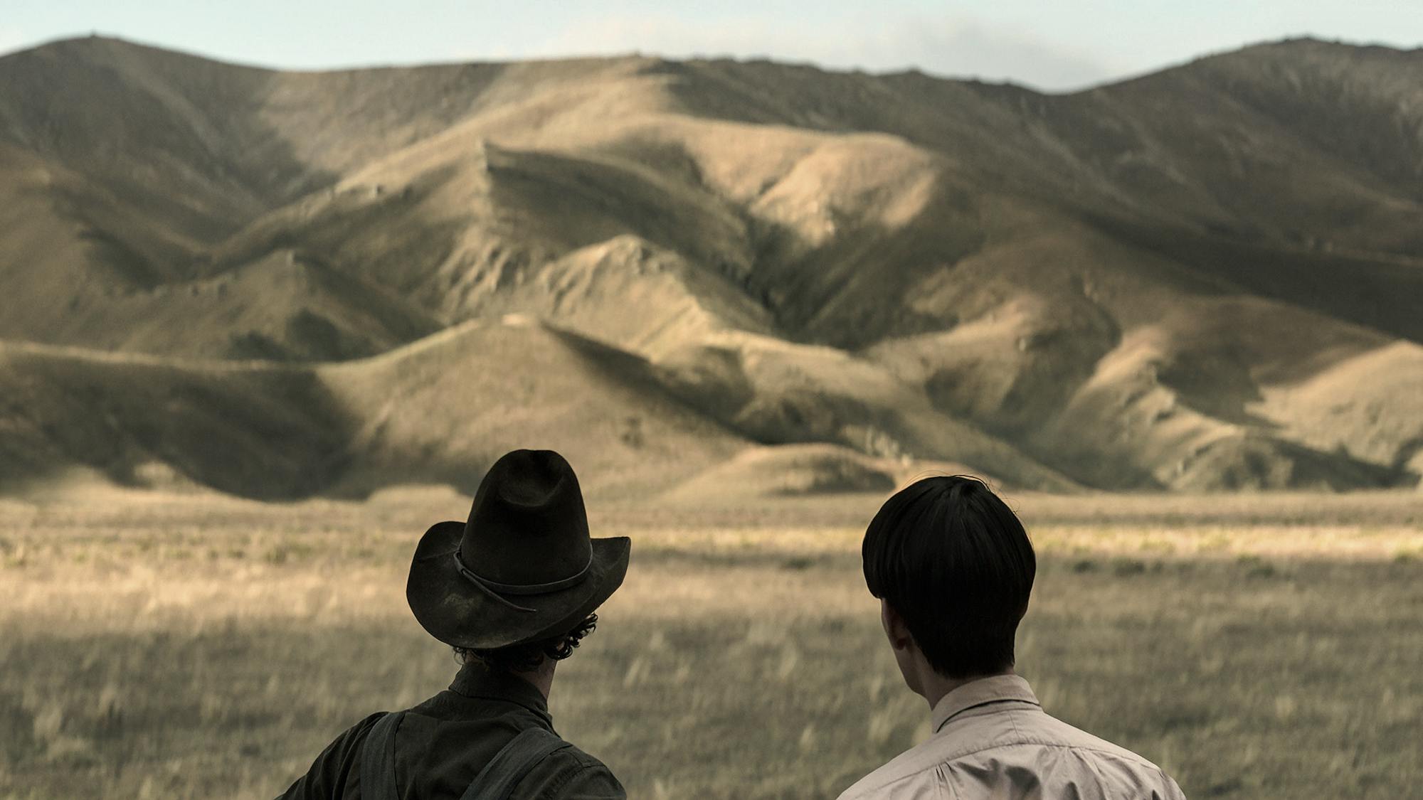 BENEDICT CUMBERBATCH as PHIL BURBANK, KODI SMIT-McPHEE as PETER in THE POWER OF THE DOG stand in front of a looming mountain range.