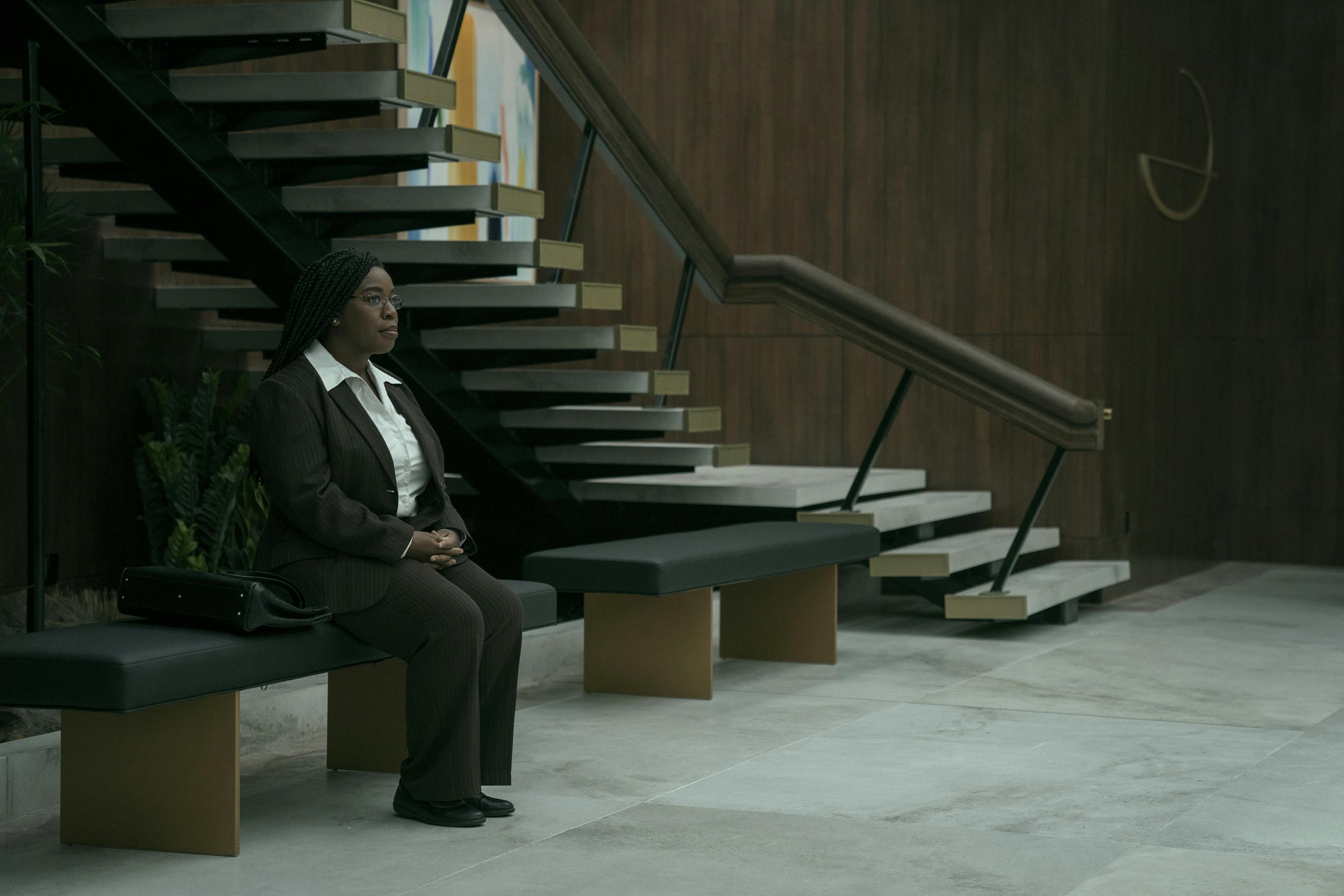 Edie Flowers (Uzo Aduba) sits on a bench. She wears a brown suit and a white shirt with a pointy collar and has extremely good posture. Behind her is a staircase that looks like it's made of Jenga blocks.