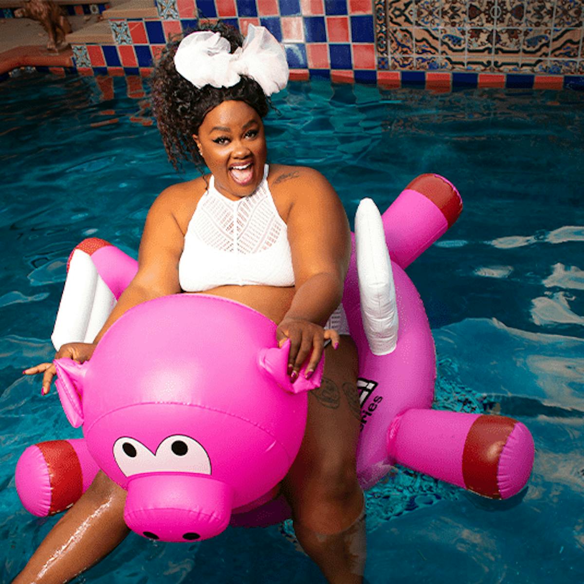 Nicole Byer sits on an inflatable pig, floating in a pool.