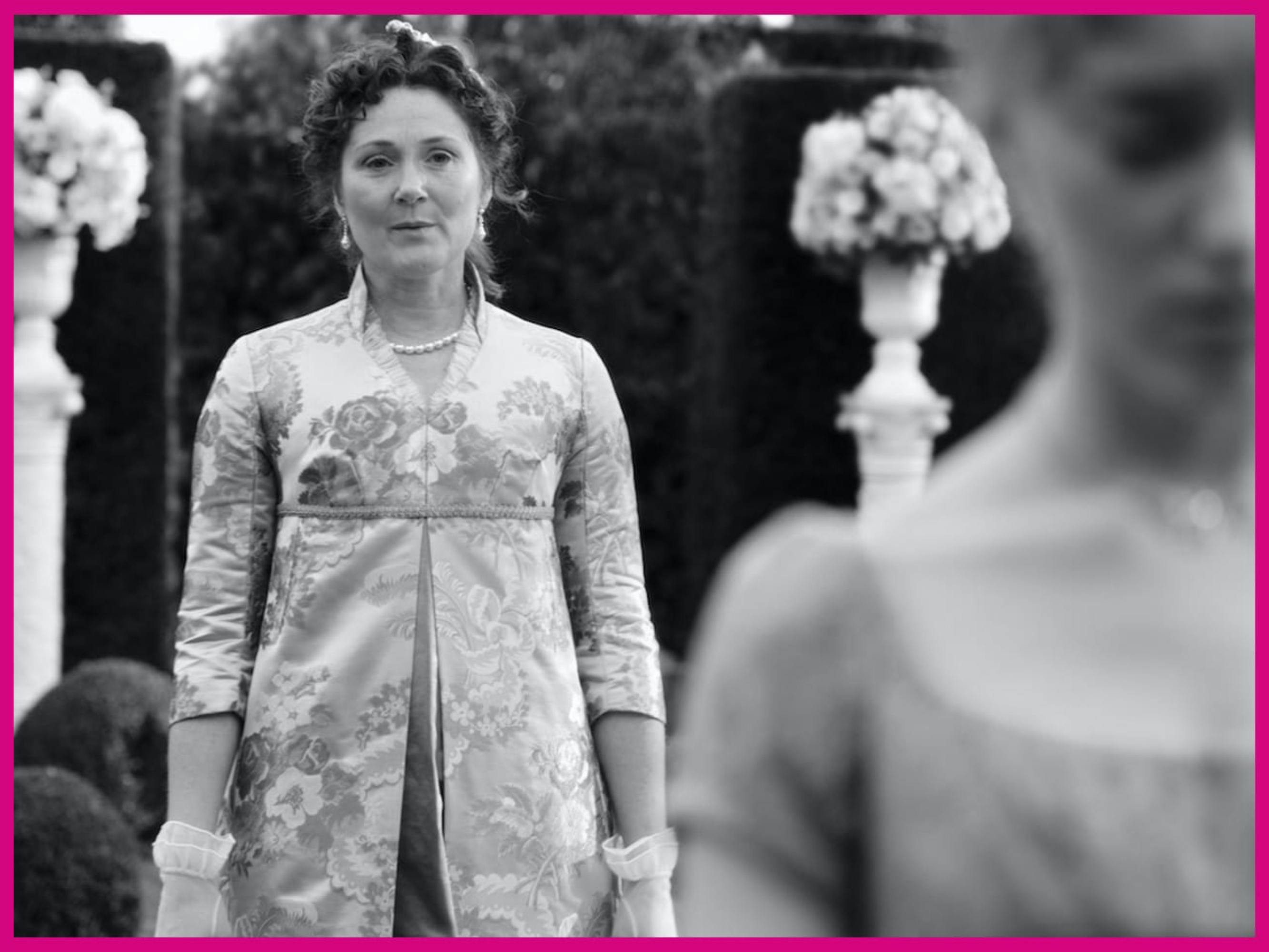 Lady Violet Bridgerton (Ruth Gemmell) and Daphne Bridgerton (Phoebe Dynevor) stand outside. Daphne walks away from her mother, with an upset expression out of focus. Violet looks concerned, and appears in focus in the center of this picture. The tense shot is juxtaposed with their light, flowery clothing, and the flowers in the background.