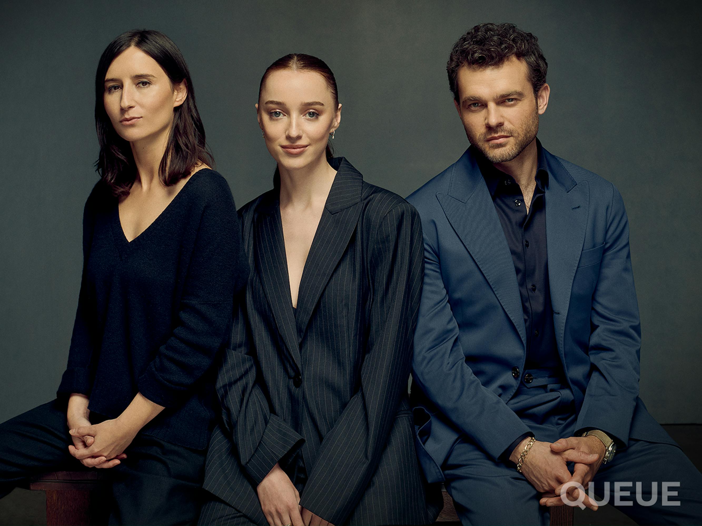 Chloe Domont, Phoebe Dynevor, and Alden Ehrenreich stand together not smiling. Chloe wears a V-neck shirt, Phoebe wears a black pinstripe suit and Alden wears a navy suit.
