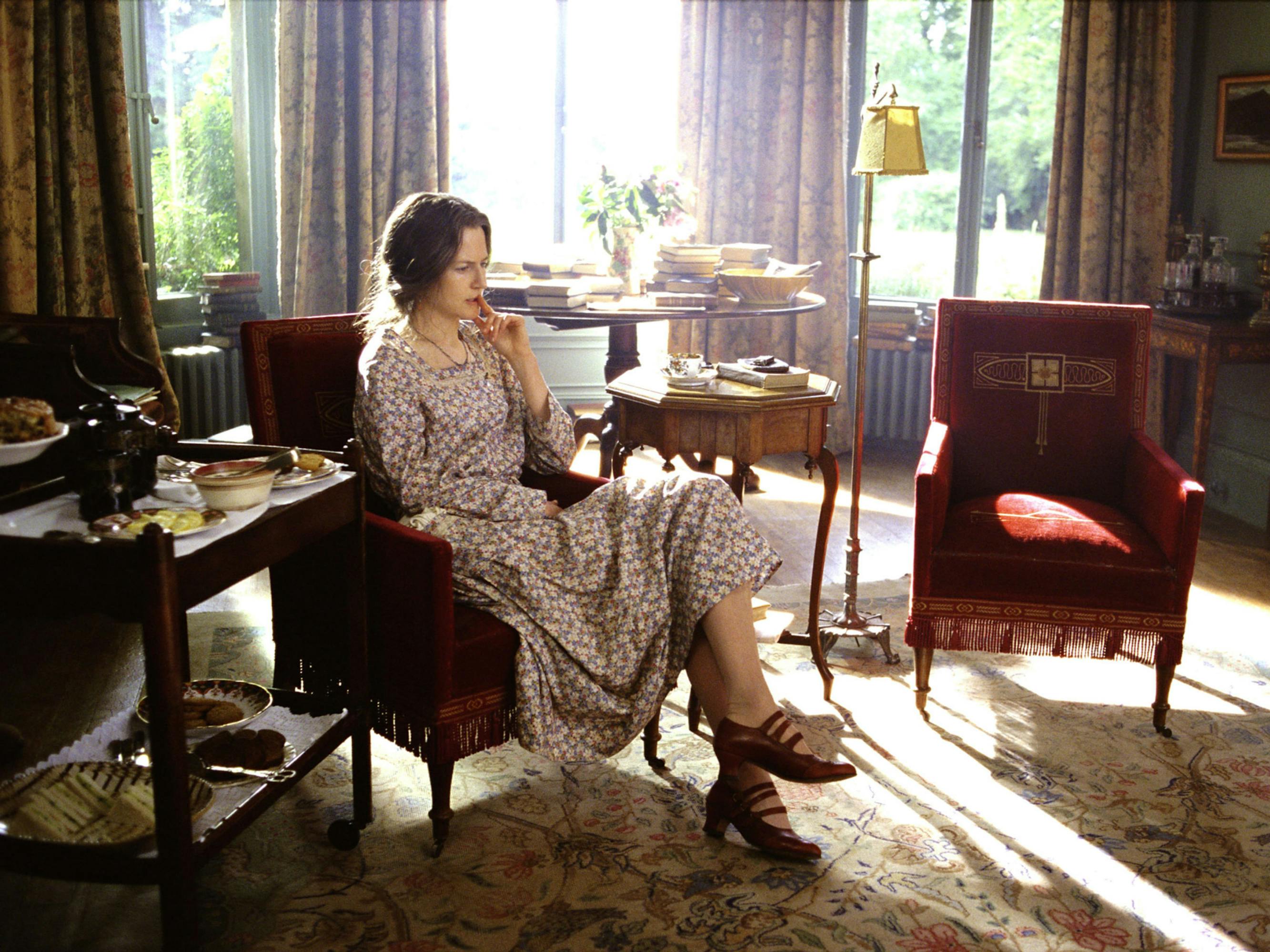 Nicole Kidman sits in a red armchair in a scene from The Hours. She wears a floral dress and low, strapped heels. 