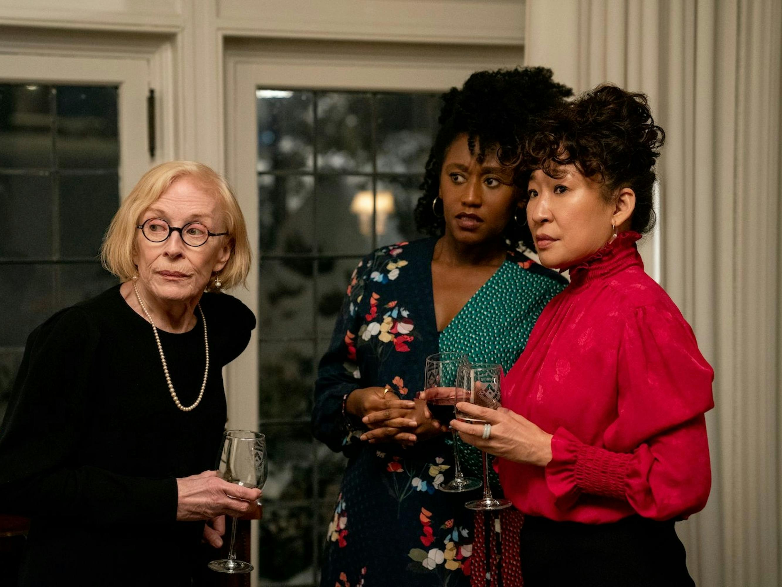 Joan Hambling (Holland Taylor), Yaz McKay (Nana Mensah), and Dr. Ji-Yoon Kim (Sandra Oh) in The Chair. The three women stand by a window discussing something off screen. Hambling wears a black outfit and glasses. McKay wears a floral top. Oh wears a red blouse and has an updo.