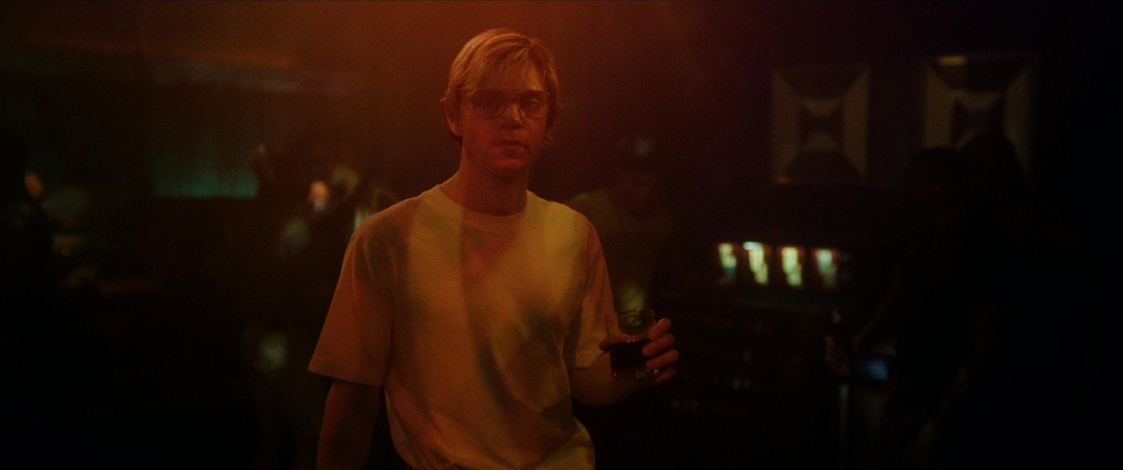 Jeffrey Dahmer (Evan Peters) wears a white t-shirt, glasses, and holds a glass of dark liquid in a smoky dark room.