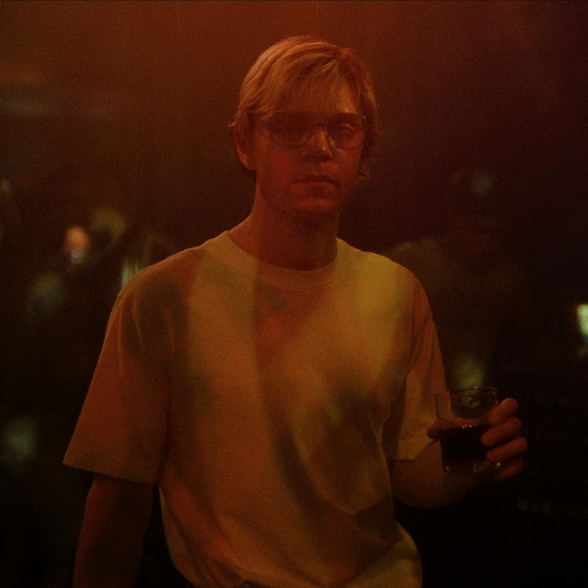Jeffrey Dahmer (Evan Peters) wears a white t-shirt, glasses, and holds a glass of dark liquid in a smoky dark room.