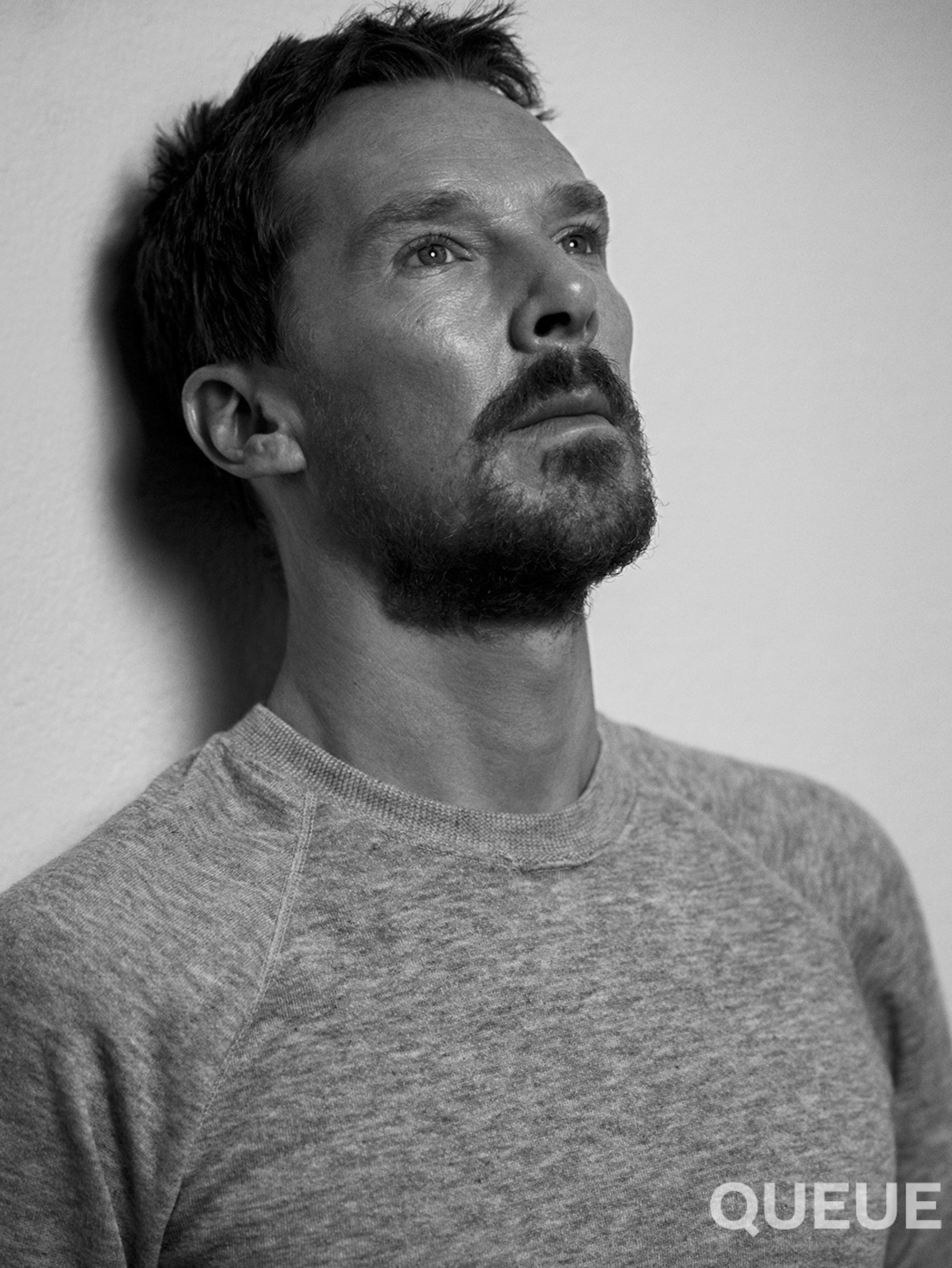 Benedict Cumberbatch wears a grey shirt and rests against a wall. He has some scruffy facial hair.