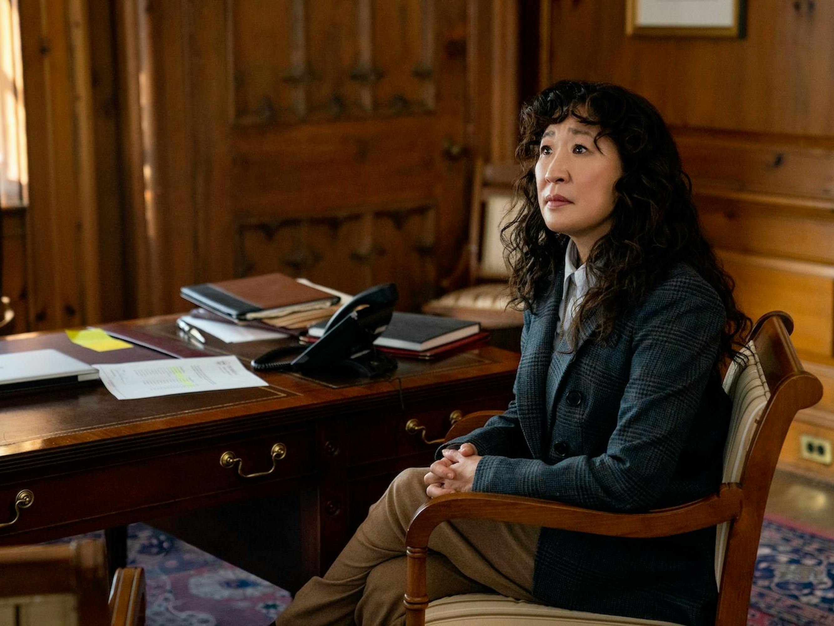 Dr. Ji-Yoon Kim (Sandra Oh) in The Chair sits in a wood paneled office wearing a black blazer. She looks attentively at someone off camera.