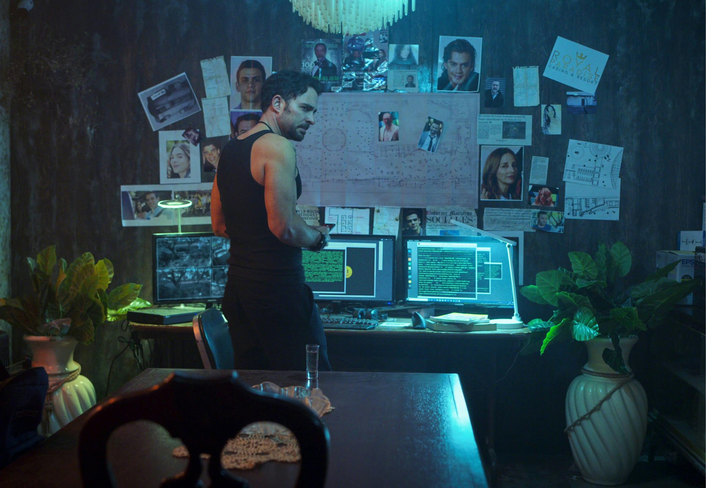 A man stands in front of a display of computers, pictures taped to the wall, and a map. He wears a black tank top and black pants, and has a sinister look on his face. There are plants flanking the table, and the room is lit with eerie blue light.