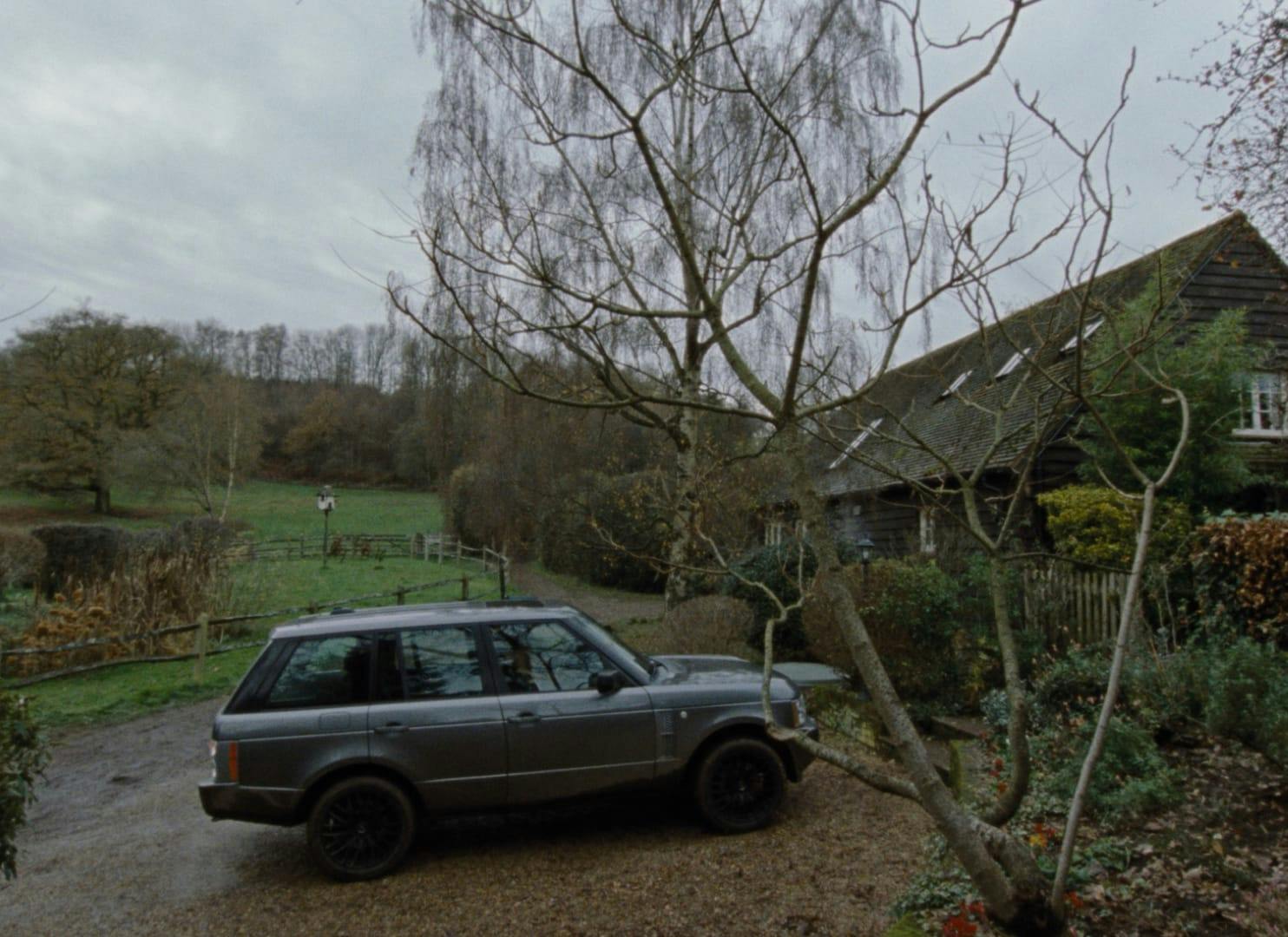 An exterior shot of the house. It is shrouded in trees and bushes, and there is a car parked in the driveway. The yard is sprawling, with fences, trees, and bushes.