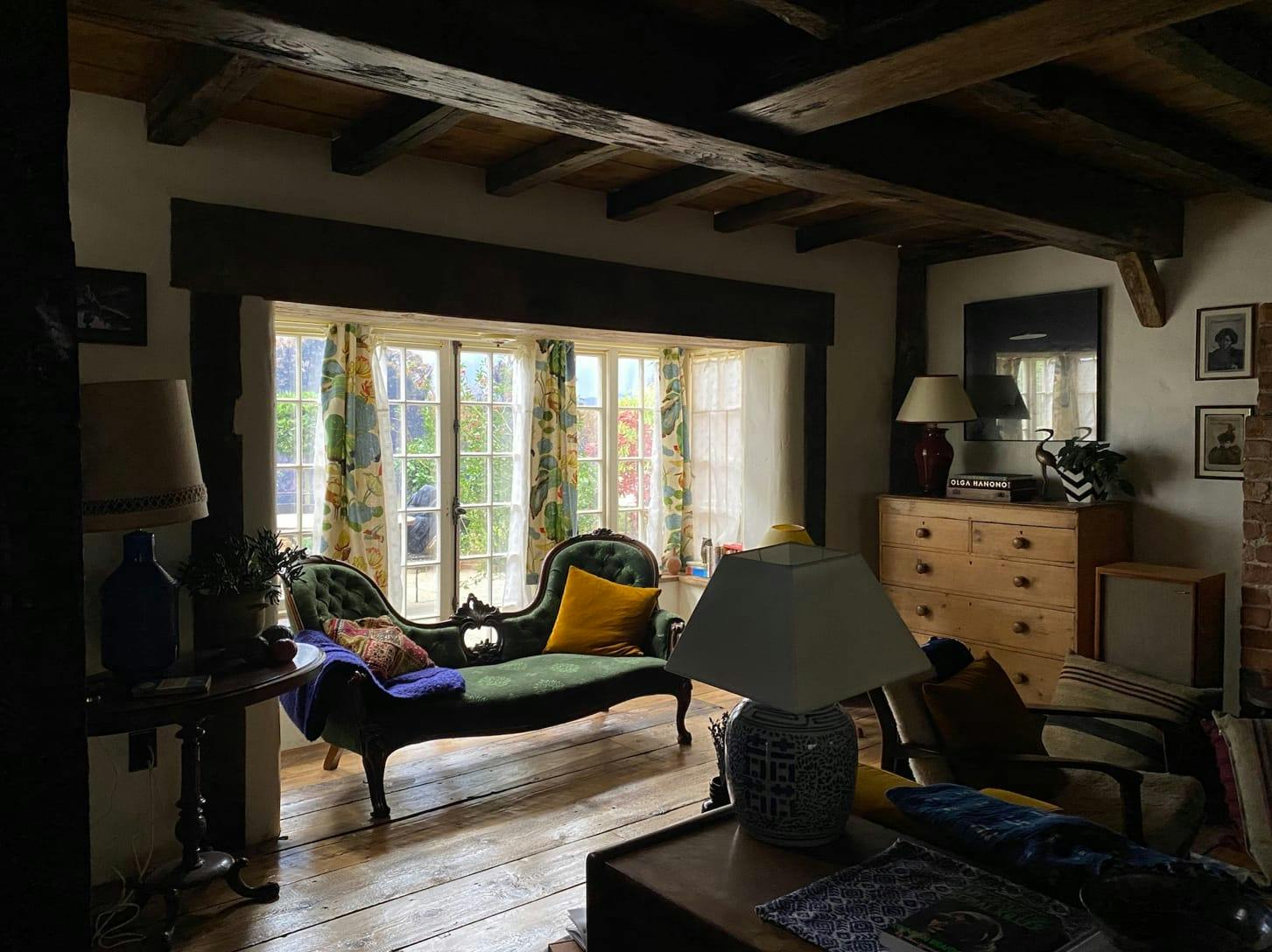 A green couch with paisley and yellow pillows, as well as a blue blanket. The french doors are open, framed by colorful curtains. In the foreground is a blue lamp.