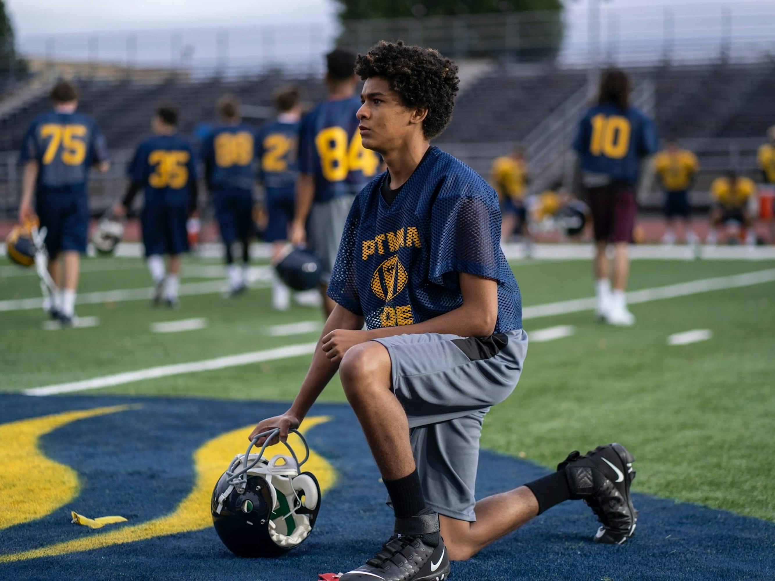 Colin Kaepernick (Jaden Michael) wears a navy, grey, and yellow uniform as he kneels on a turf field.