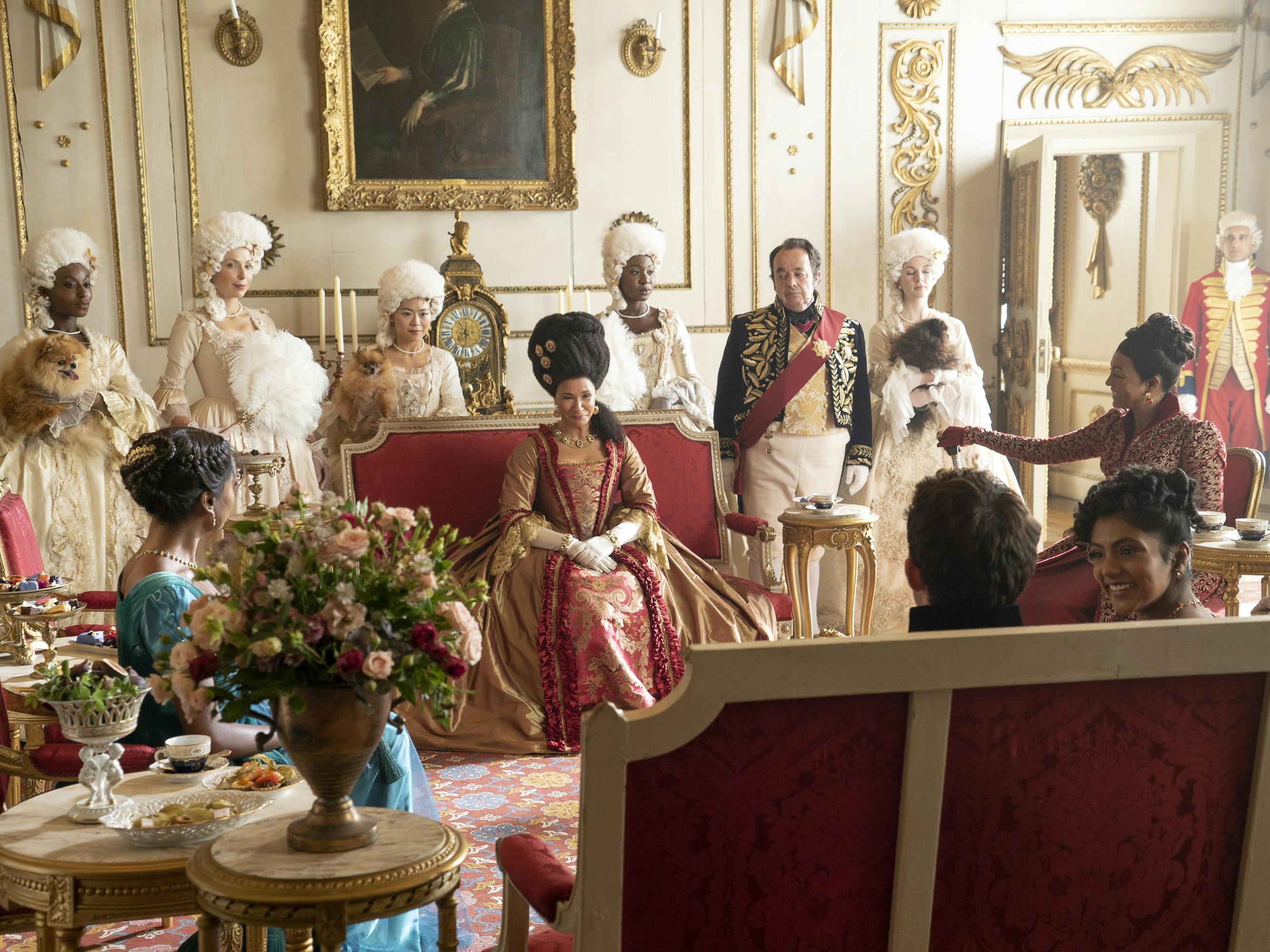 Kate Sharma (Simone Ashley), Queen Charlotte (Golda Rosheuvel), Lady Danbury (Adjoa Andoh), Edwina Sharma (Charithra Chandran), and Anthony Bridgerton (Jonathan Bailey) sit around an ornate living room surrounded by people in wigs.