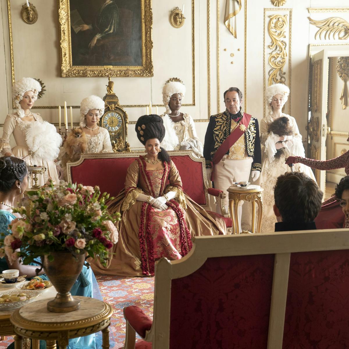 Kate Sharma (Simone Ashley), Queen Charlotte (Golda Rosheuvel), Lady Danbury (Adjoa Andoh), Edwina Sharma (Charithra Chandran), and Anthony Bridgerton (Jonathan Bailey) sit around an ornate living room surrounded by people in wigs.