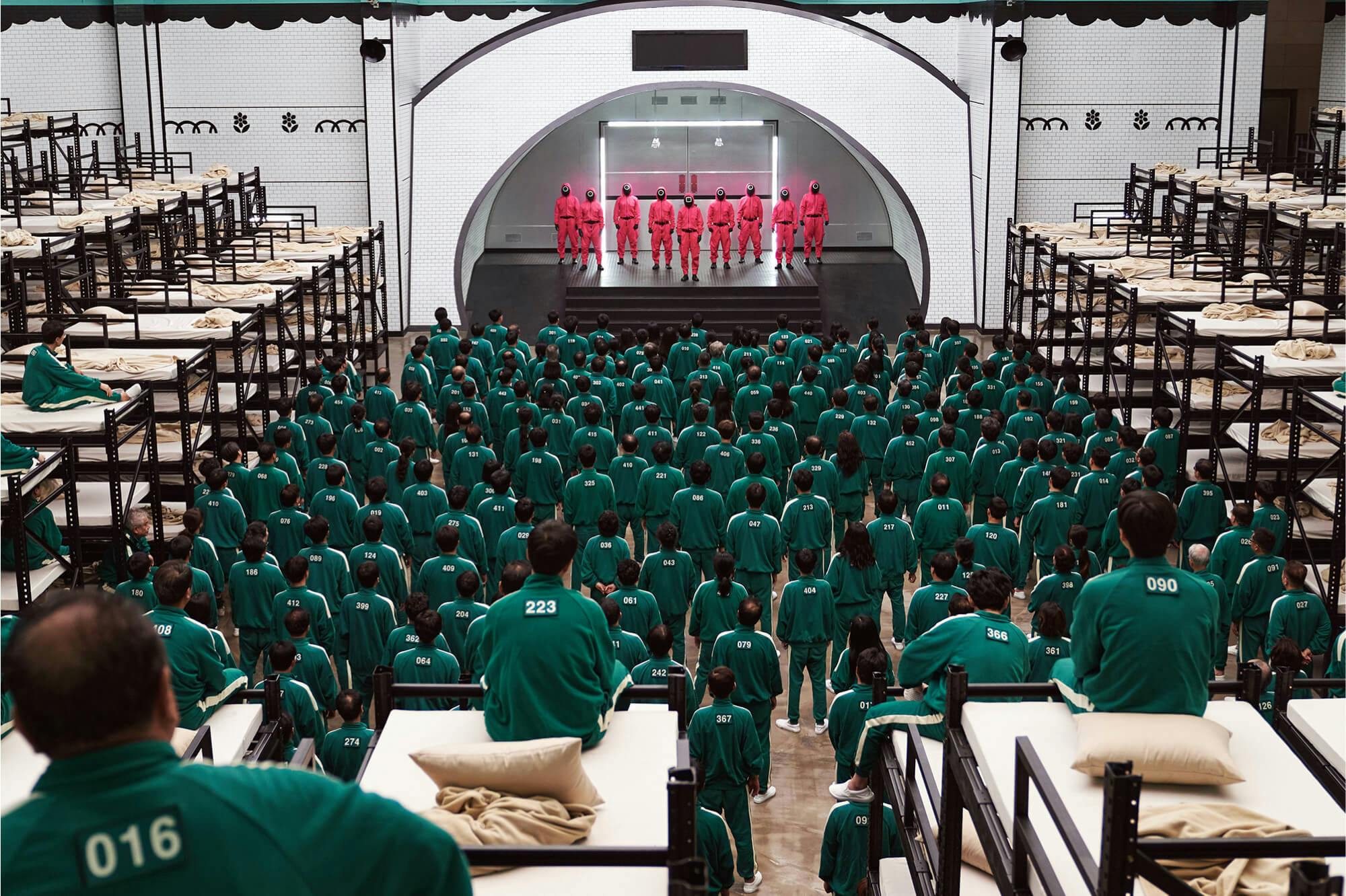 A shot of one of the halls in Squid Game. 456 players line up in green tracksuits labeled with their numbers, facing a stage of red-clothed people wearing black masks.
