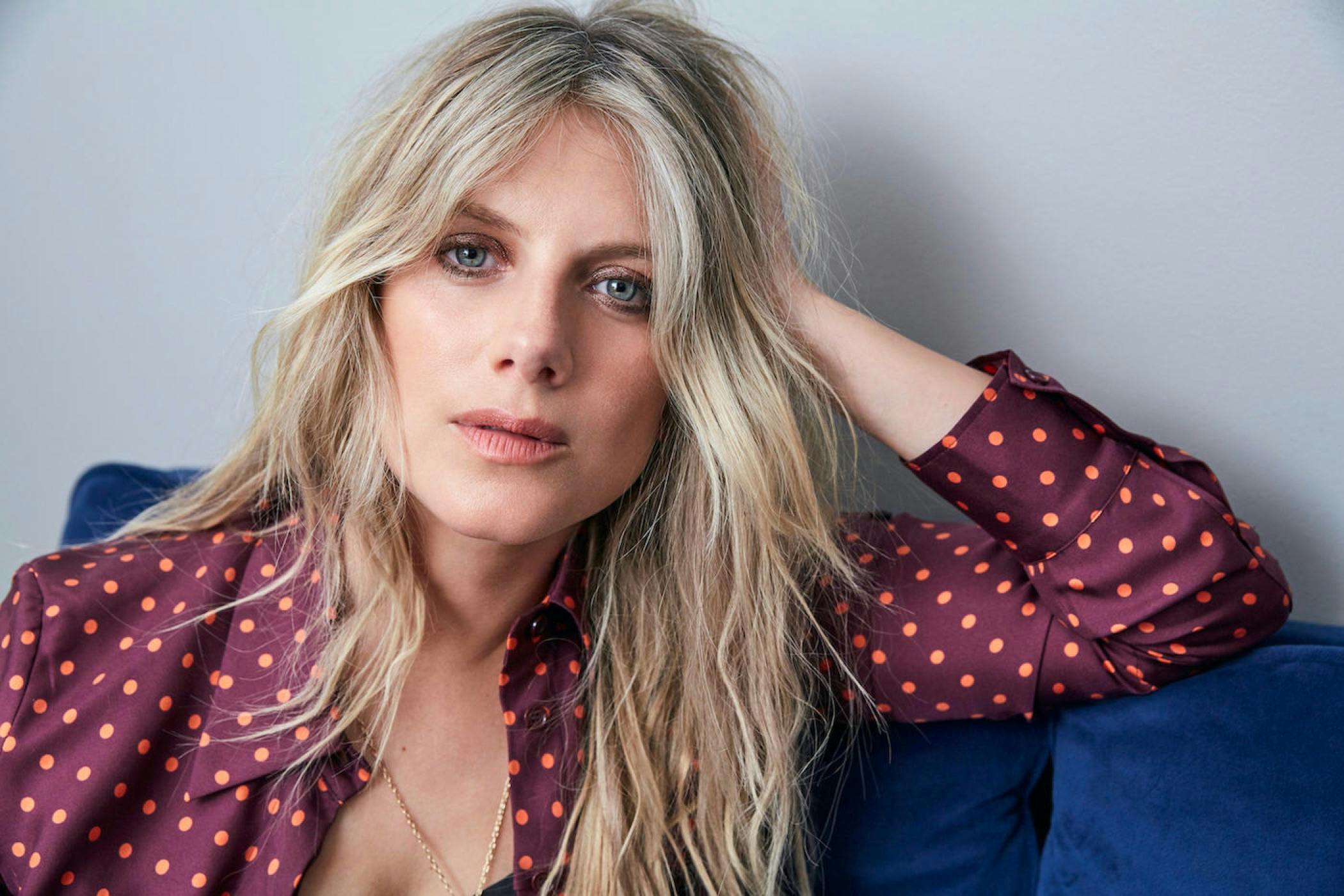 Laurent wears a maroon and orange-polka-dotted blouse, gold jewelry, and sits on a blue couch. Her blond hair is long, and she stares right at the camera.