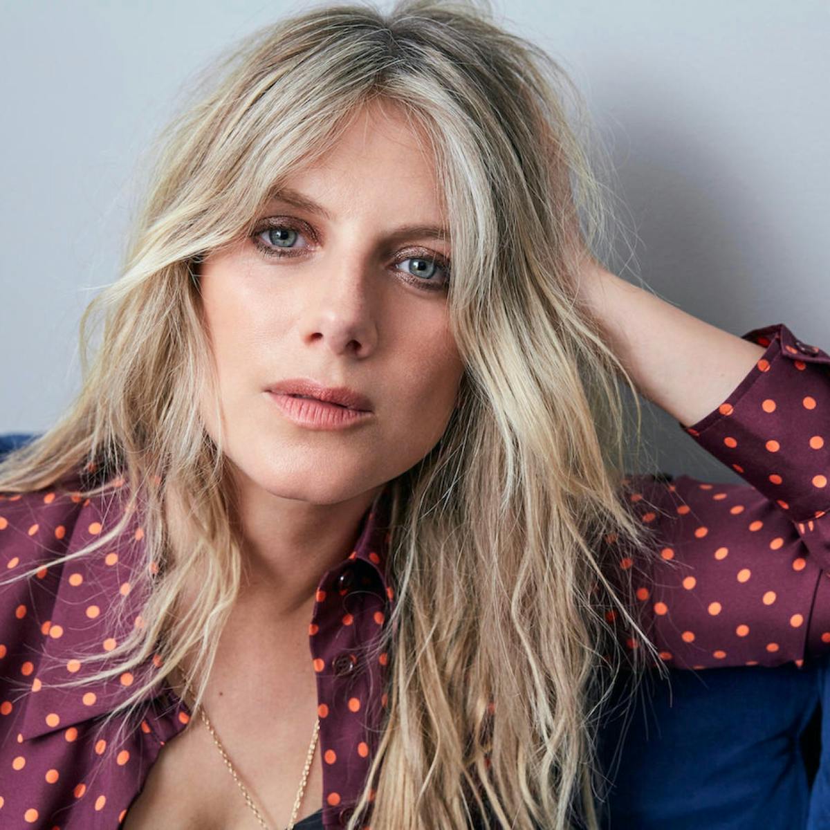 Laurent wears a maroon and orange-polka-dotted blouse, gold jewelry, and sits on a blue couch. Her blond hair is long, and she stares right at the camera.