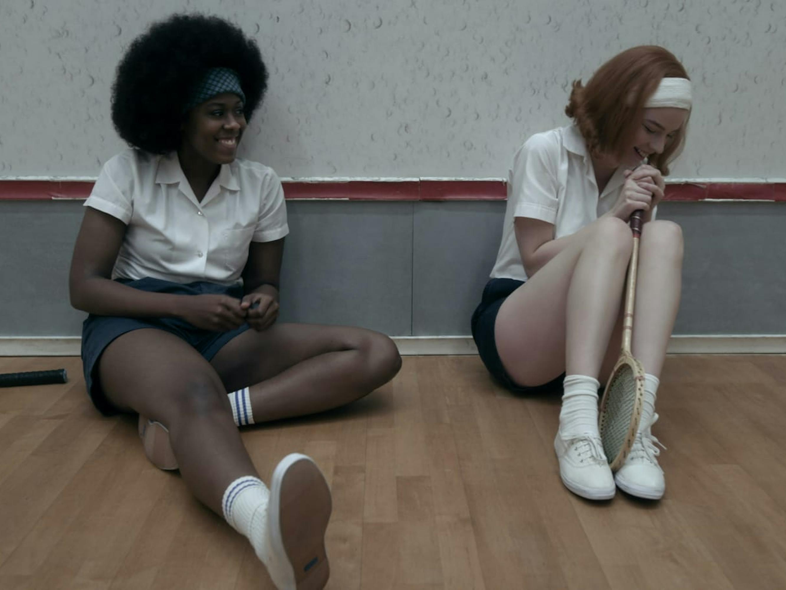 Jolene (Moses Ingram) and Beth Harmon (Anya Taylor-Joy) sit against a squash court wall. Both girls sport white collared shirts, dark shorts, headbands, white keds, and white socks. Taylor-Joy holds her racket between her legs, while Jolene’s lies on the ground beside her.