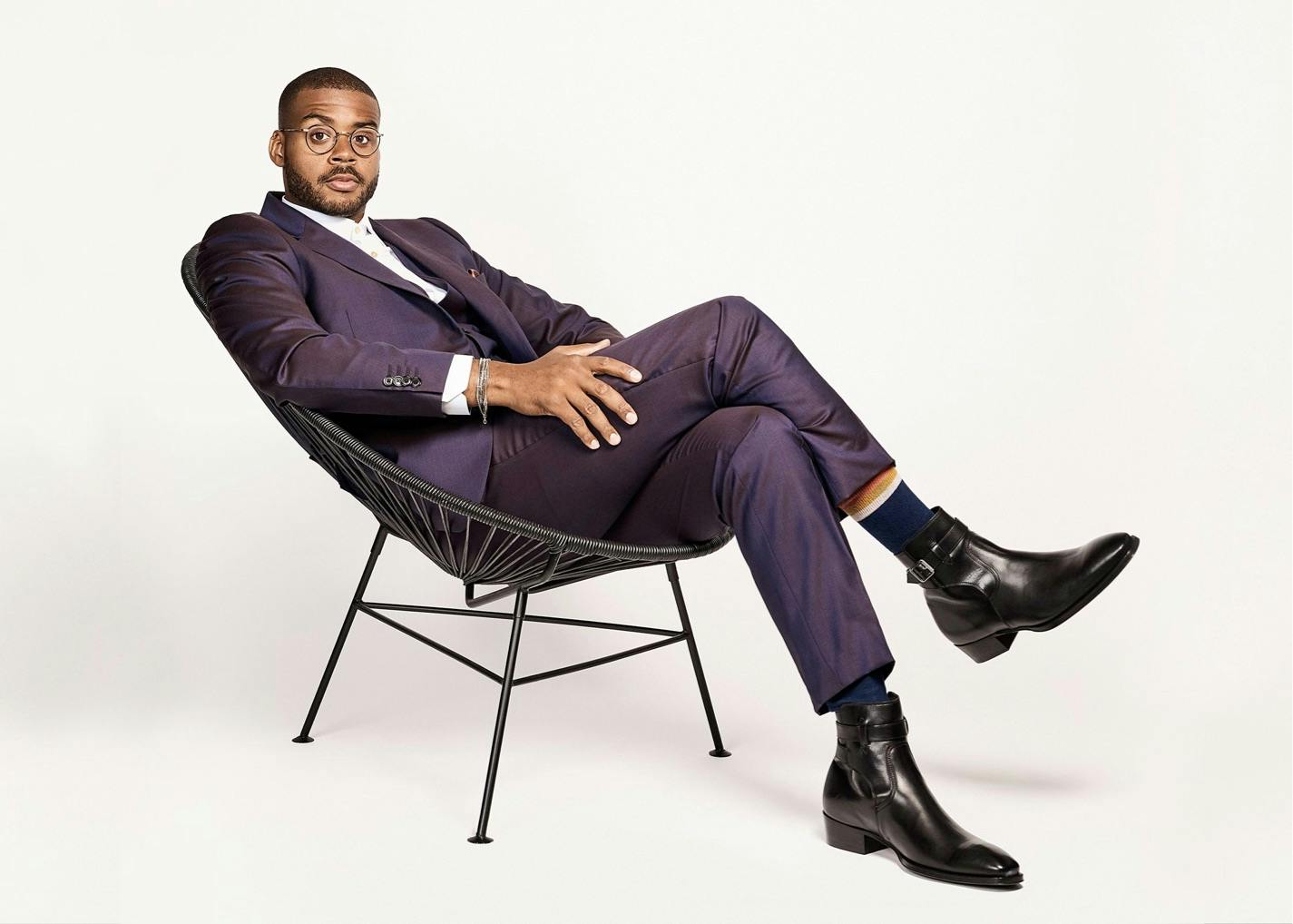 Kris Bowers sits in a black chair. He wears a beautiful purplish-blue suit, white shirt, black boots, navy socks, and circular glasses. He looks chic and calm as he looks directly at the camera.