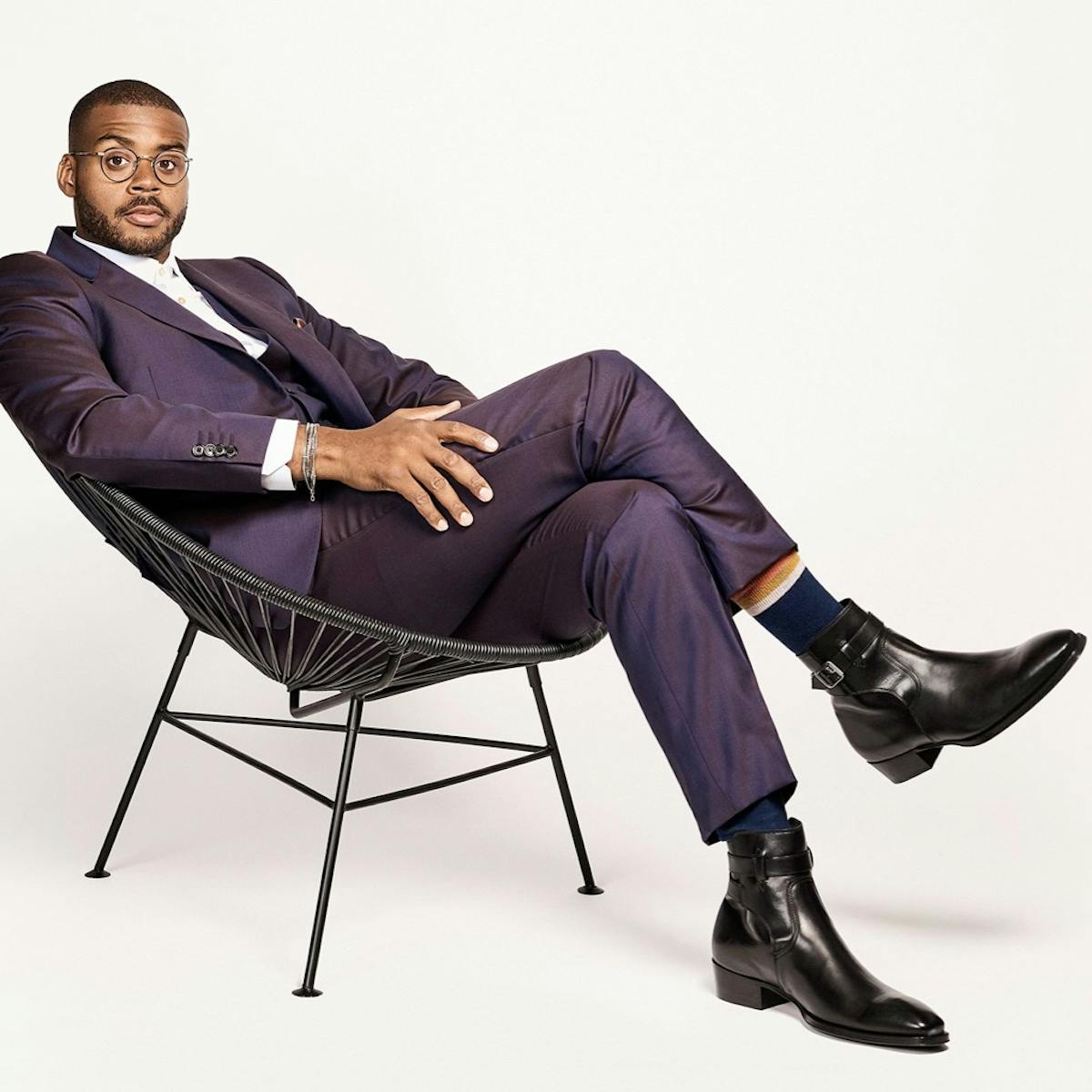 Kris Bowers sits in a black chair. He wears a beautiful purplish-blue suit, white shirt, black boots, navy socks, and circular glasses. He looks chic and calm as he looks directly at the camera.
