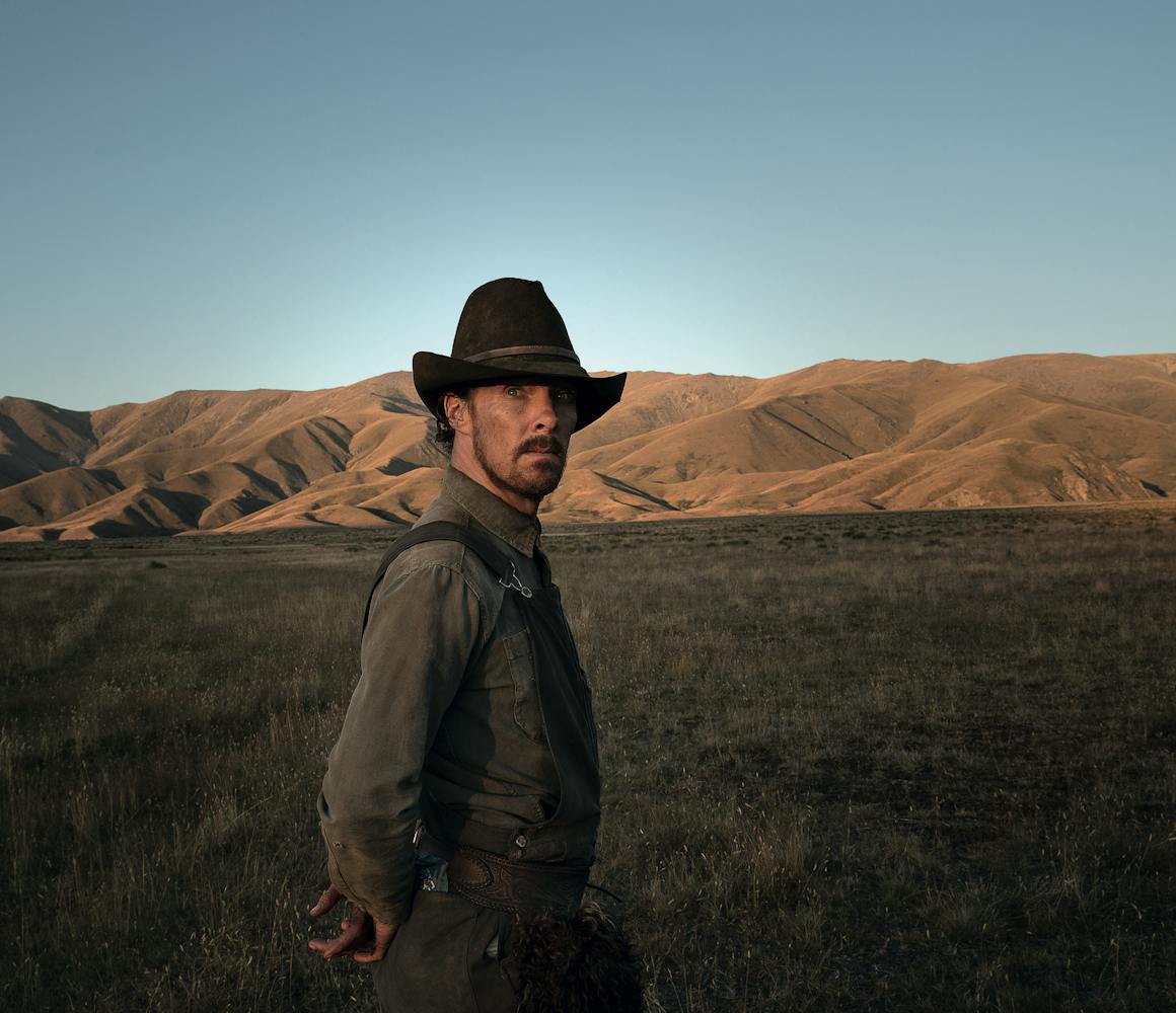 Benedict Cumberbatch wears overalls, a grey shirt, and a brown wide-brimmed hat. Behind him are sunlit mountains.
