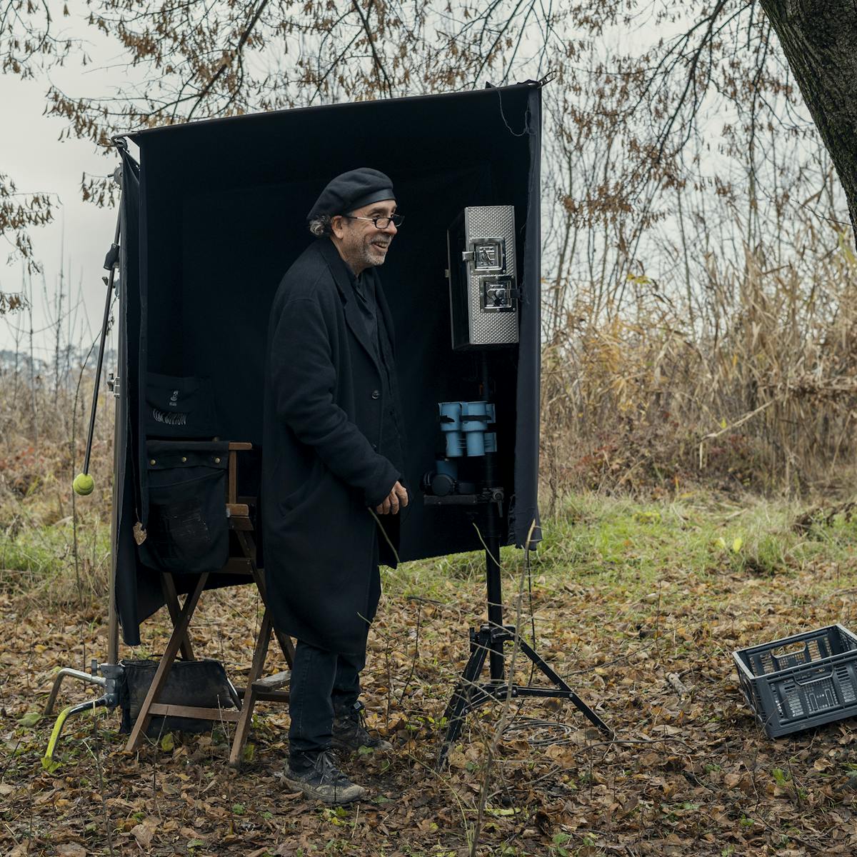 Tim Burton wears all black and stands in an autumnal setting next to some filming equipment. 