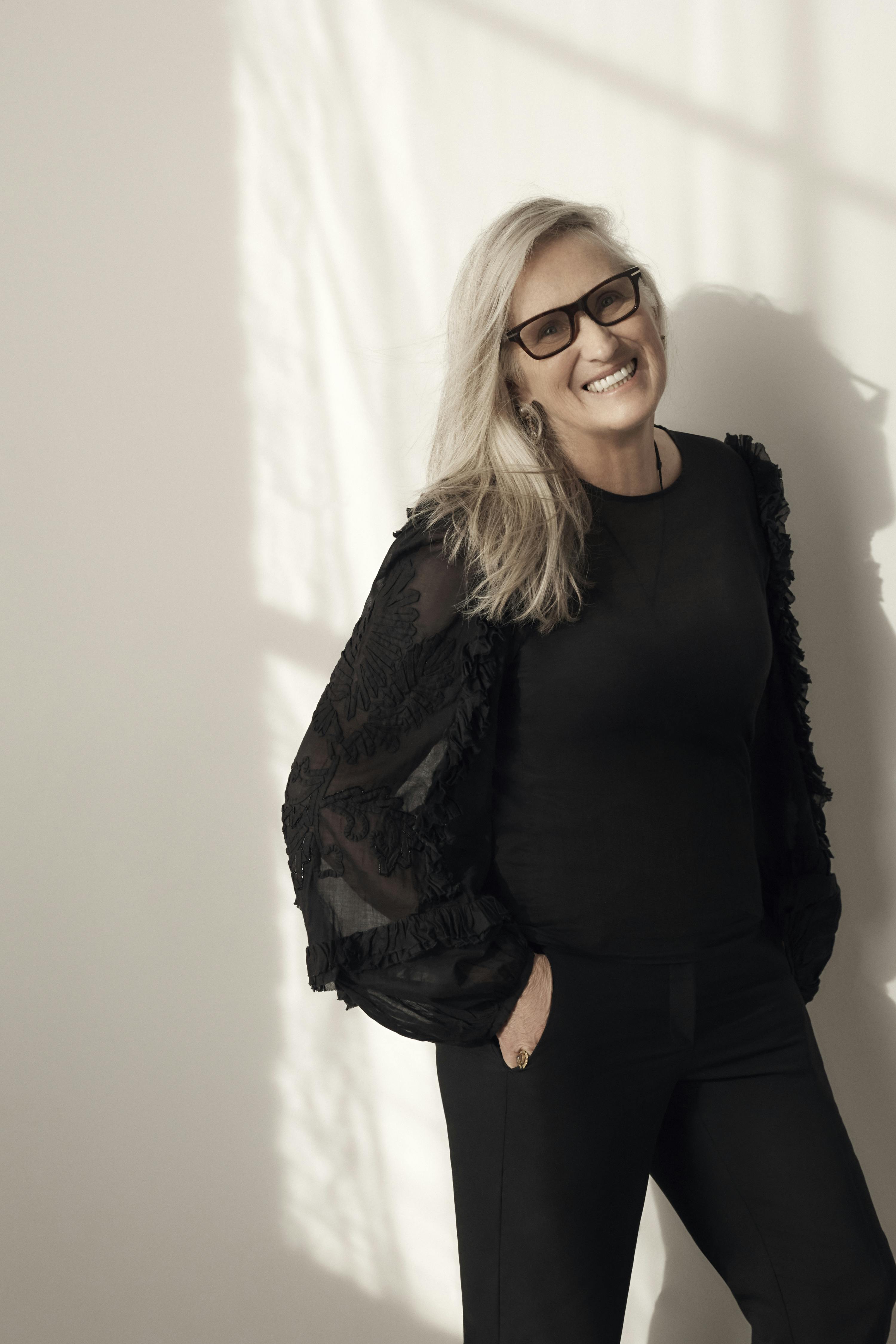 Jane Campion wears an all-black outfit and chunky black glasses. She leans again a white wall.