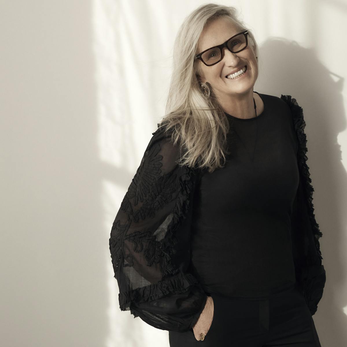 Jane Campion wears an all-black outfit and chunky black glasses. She leans again a white wall.