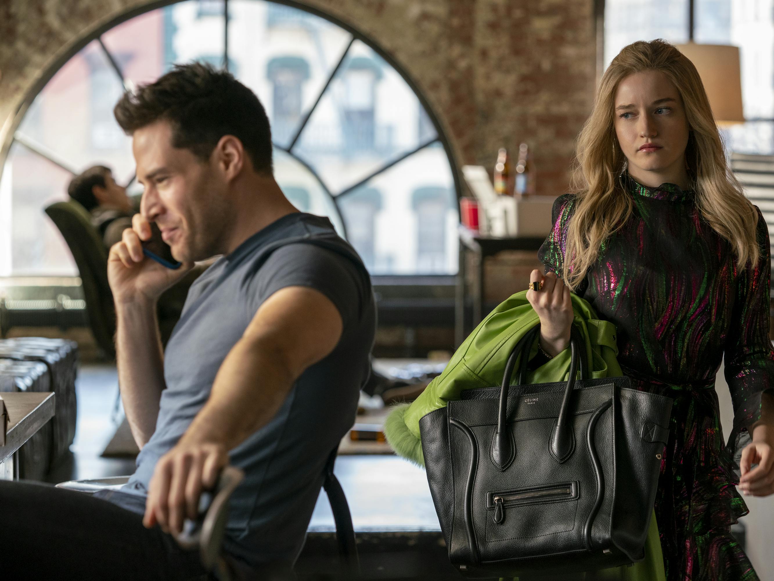 Ben Rappaport and Julia Garner. Rappaport wears a grey t-shirt and talks on the phone with a smug expression. Garner carries a black Celine back and looks disgusted.