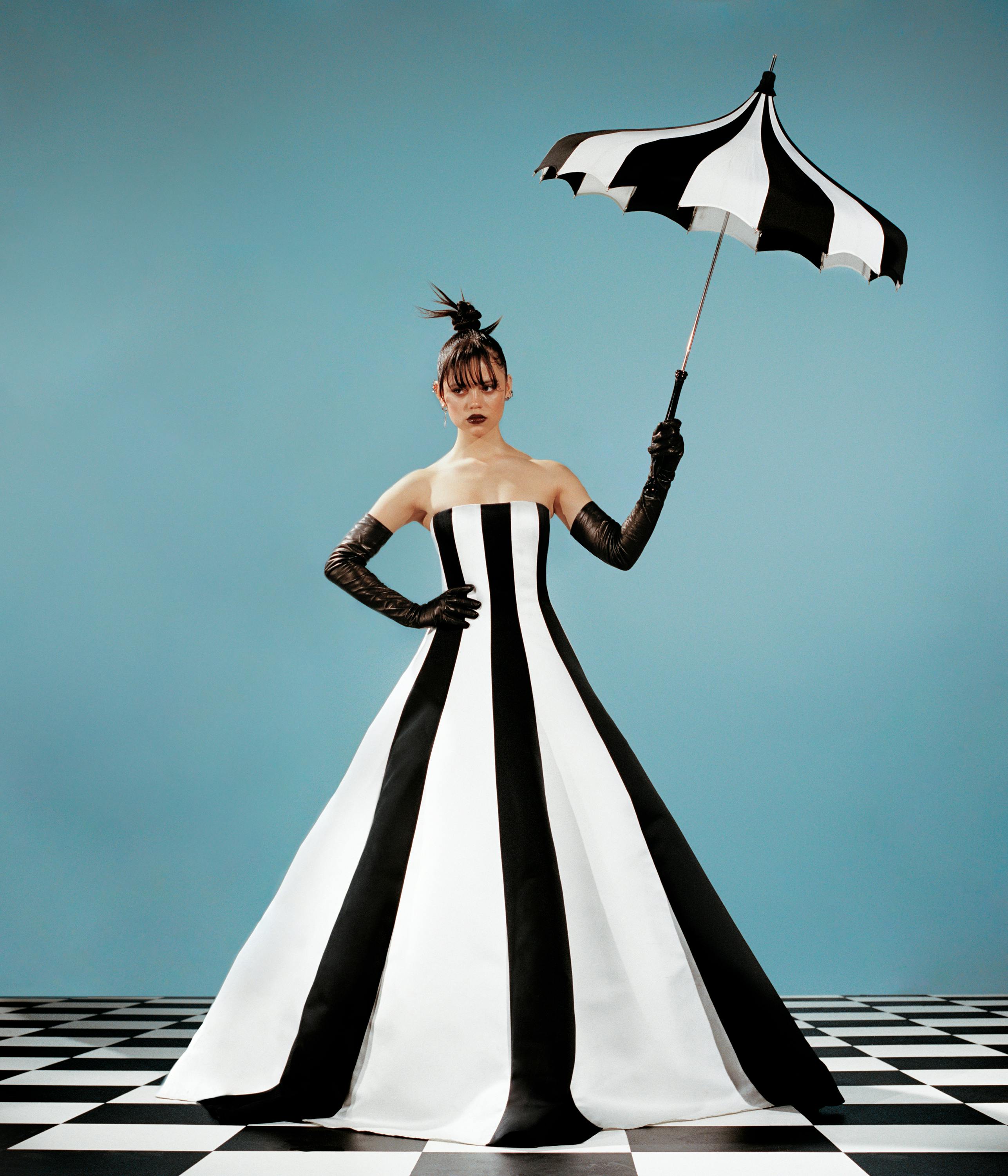 Jenna Ortega wears a black-and-white striped dress, black gloves, and carries a black-and-white umbrella. She stands on a black-and-white checkered floor against a teal background. 