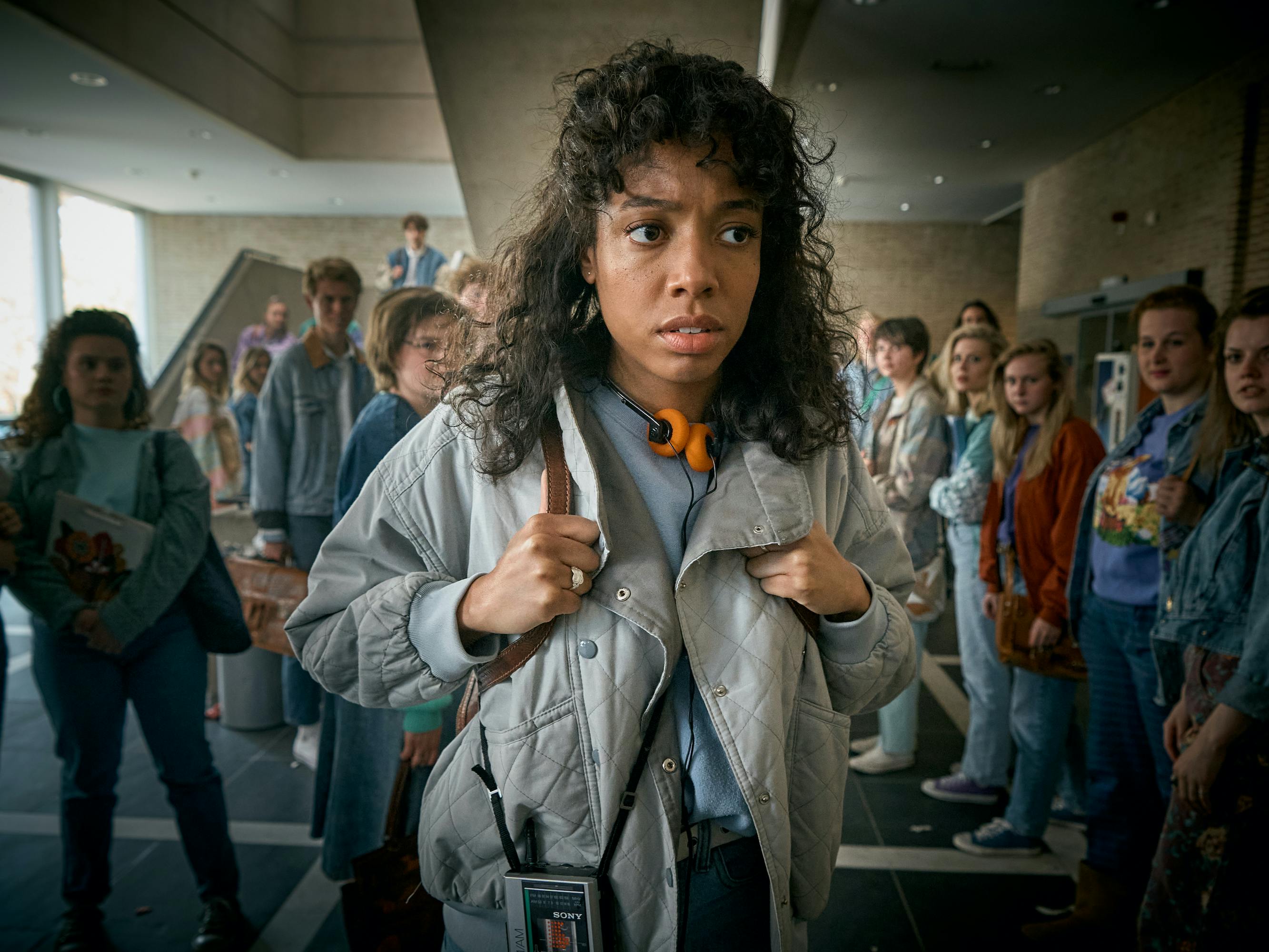 Joy Delima wears a baggy 80s jacket, and a walkman, as she walks through a crowd of staring people.