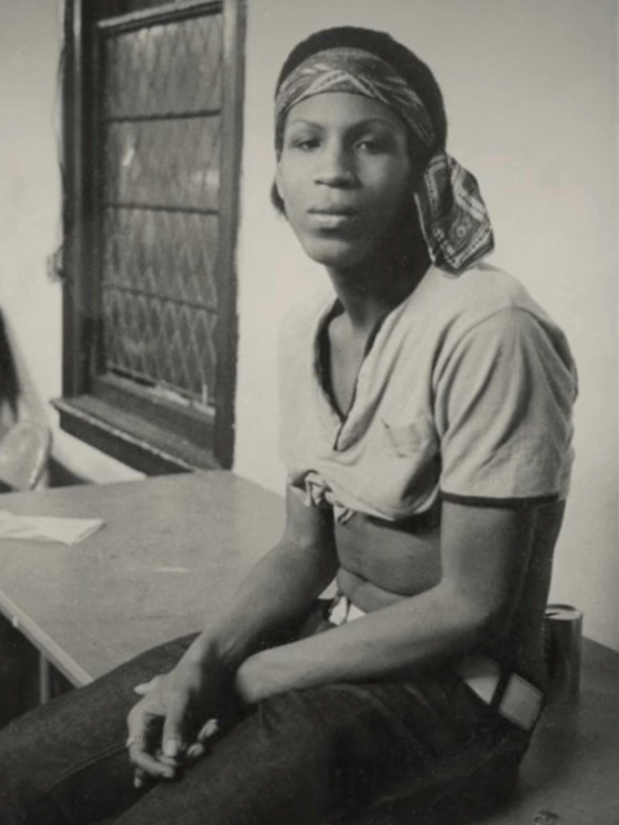 An archival, sepia-toned photo of Zazu Nova perched on a desk, looking serious and casually stylish in a cropped t-shirt and bandana.