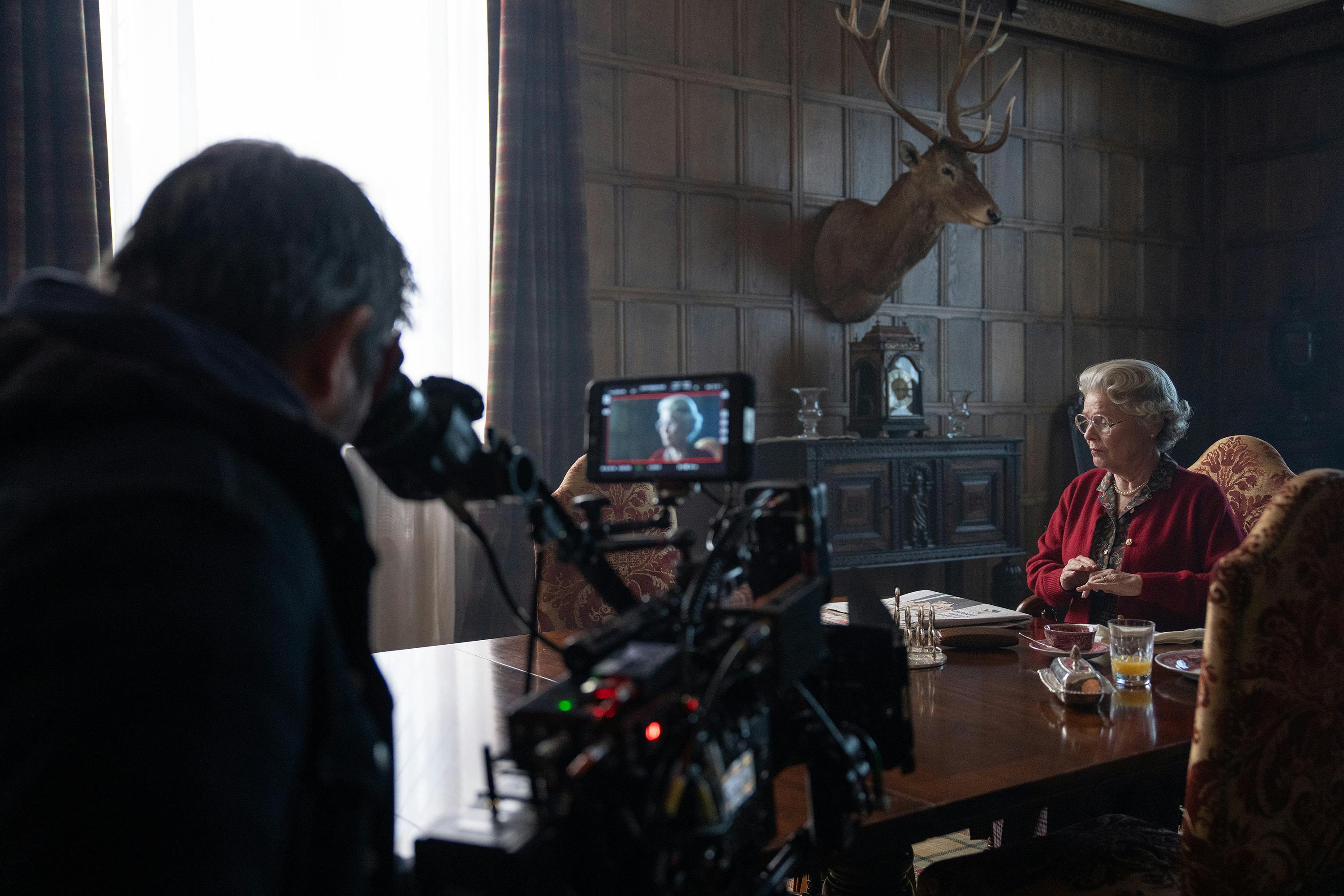 Behind the scenes of The Crown. A man stands behind a camera and shoots Imelda Staunton, who wears a red sweater and sits at a wooden table.