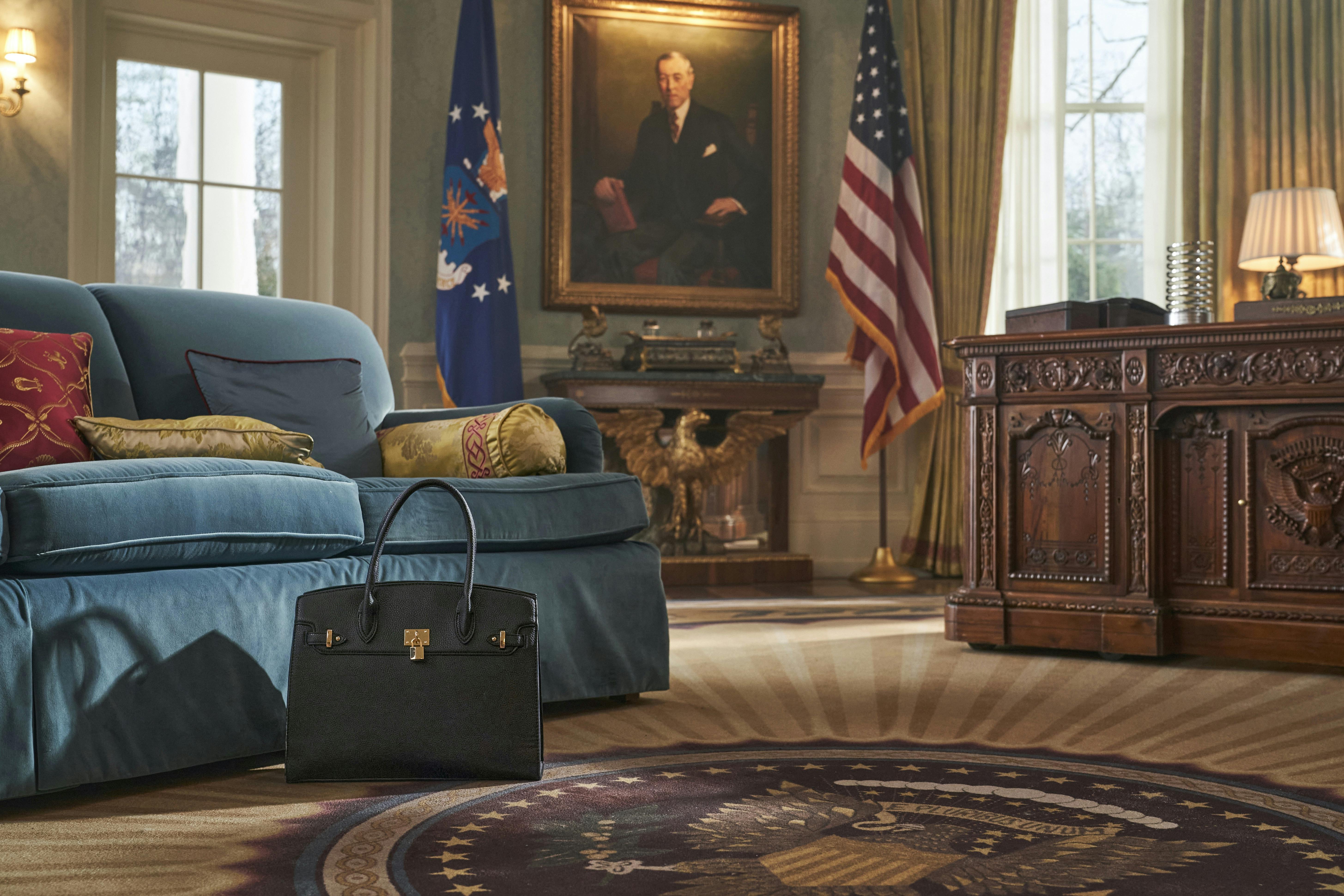 A black Birkin sits next to a blue couch. The floor is emblazoned with an oval office crest, on the wall are two flags and a portrait. There is also an old wooden desk with a lamp. 