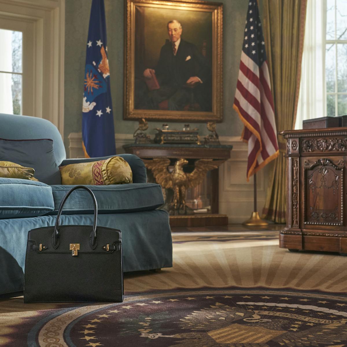 A black Birkin sits next to a blue couch. The floor is emblazoned with an oval office crest, on the wall are two flags and a portrait. There is also an old wooden desk with a lamp. 